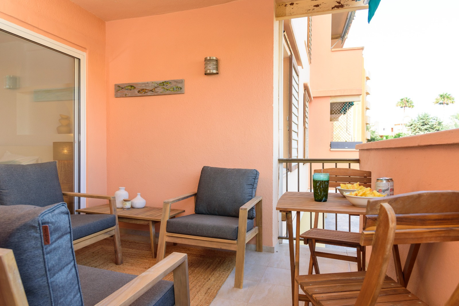 Terraza privada con paredes en tono salmón, equipada con mobiliario de madera, cojines grises y zona de comedor exterior. Incluye acceso directo al interior mediante ventanal.