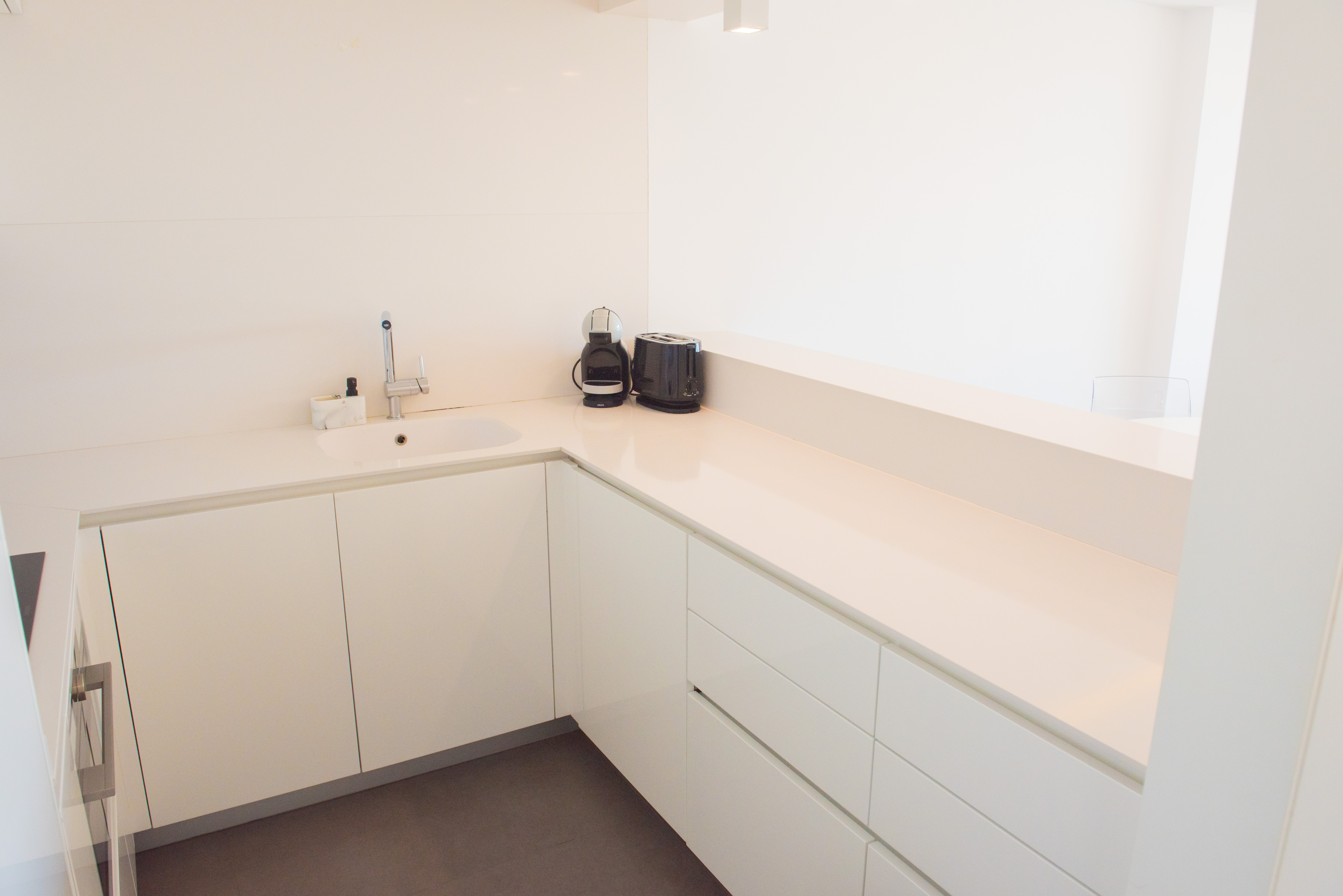 Cocina moderna de diseño minimalista en blanco brillante con encimera de cuarzo, fregadero integrado y suelo de gres en tono oscuro.