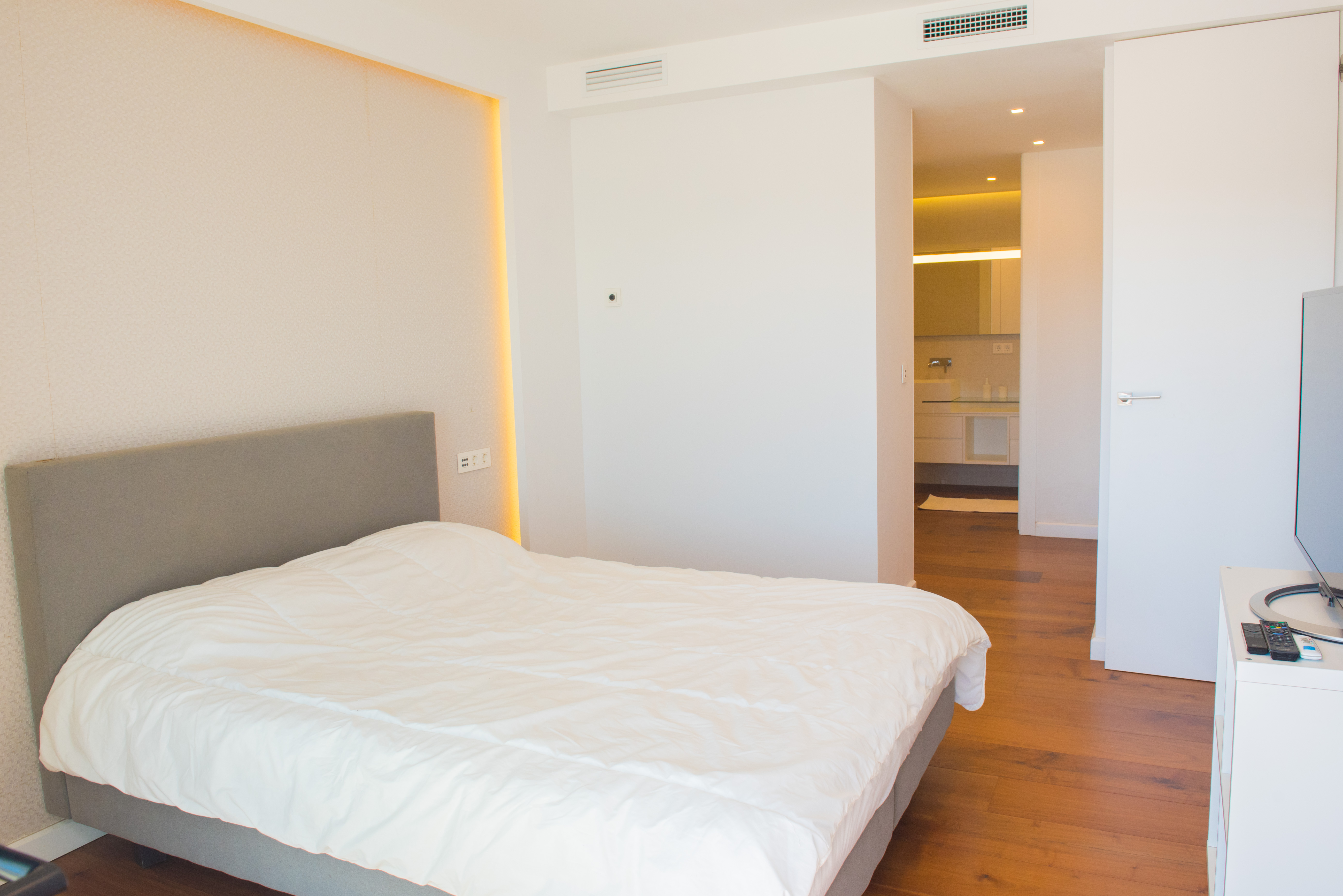 Dormitorio principal con cabecero tapizado en gris, pared con iluminación LED indirecta, suelos de madera y baño en suite integrado con acabados modernos.