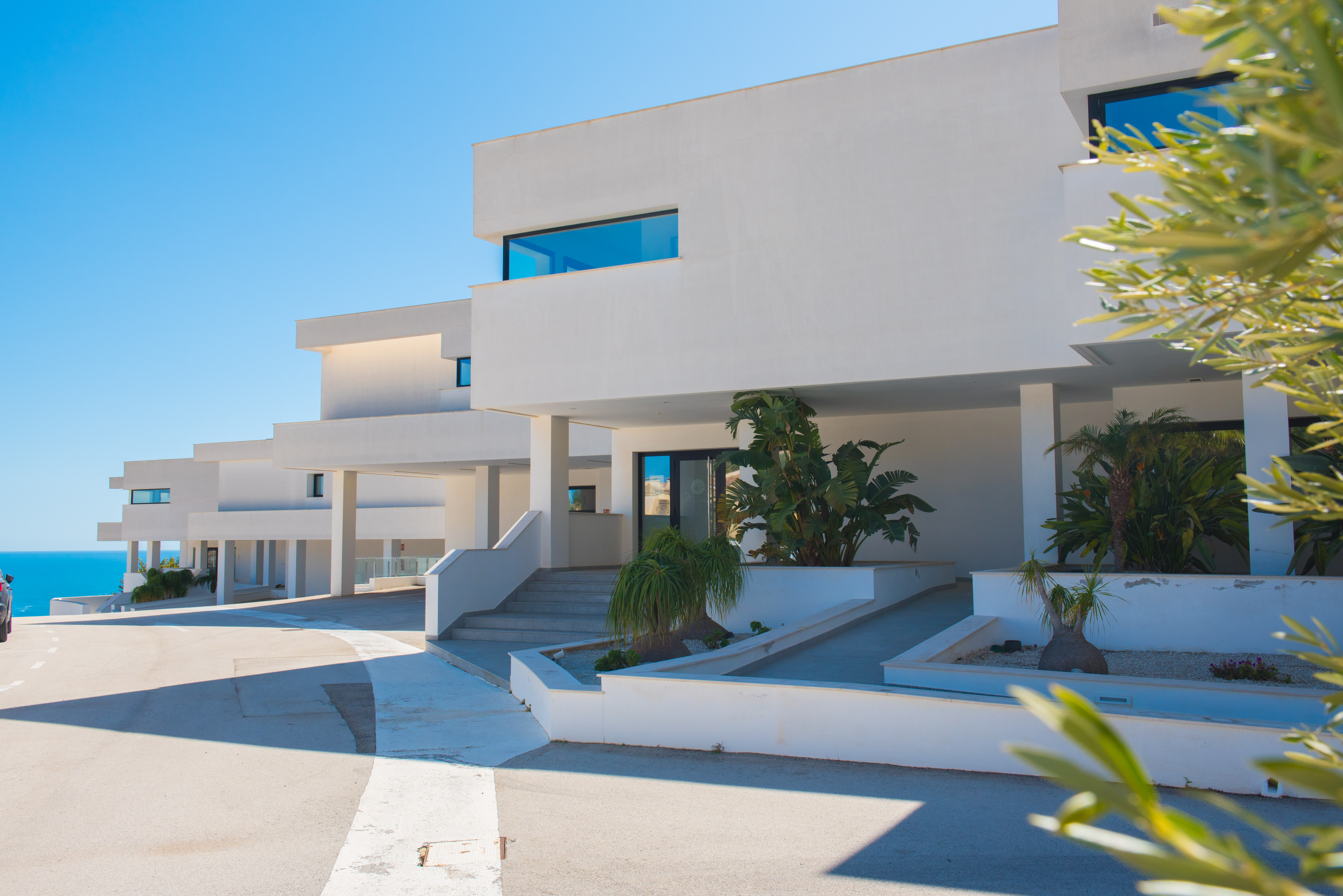 Fachada minimalista de color blanco puro con accesos escalonados y rampas, integrando jardineras con vegetación tropical y líneas arquitectónicas limpias.