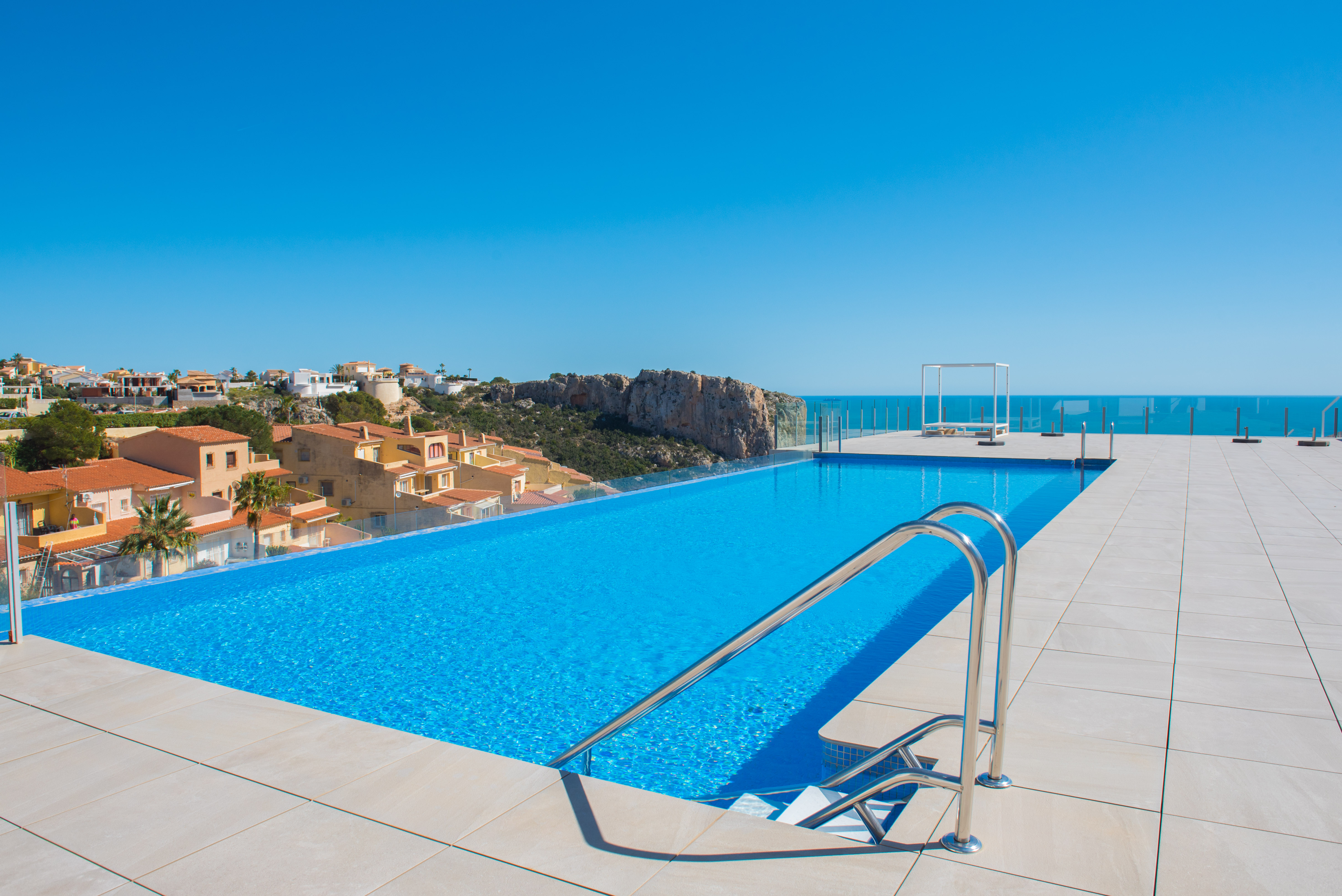 Piscina infinita comunitaria con barandillas de cristal y vistas panorámicas al mar y acantilados. El solárium está pavimentado con gres cerámico de gran formato en tonos claros.