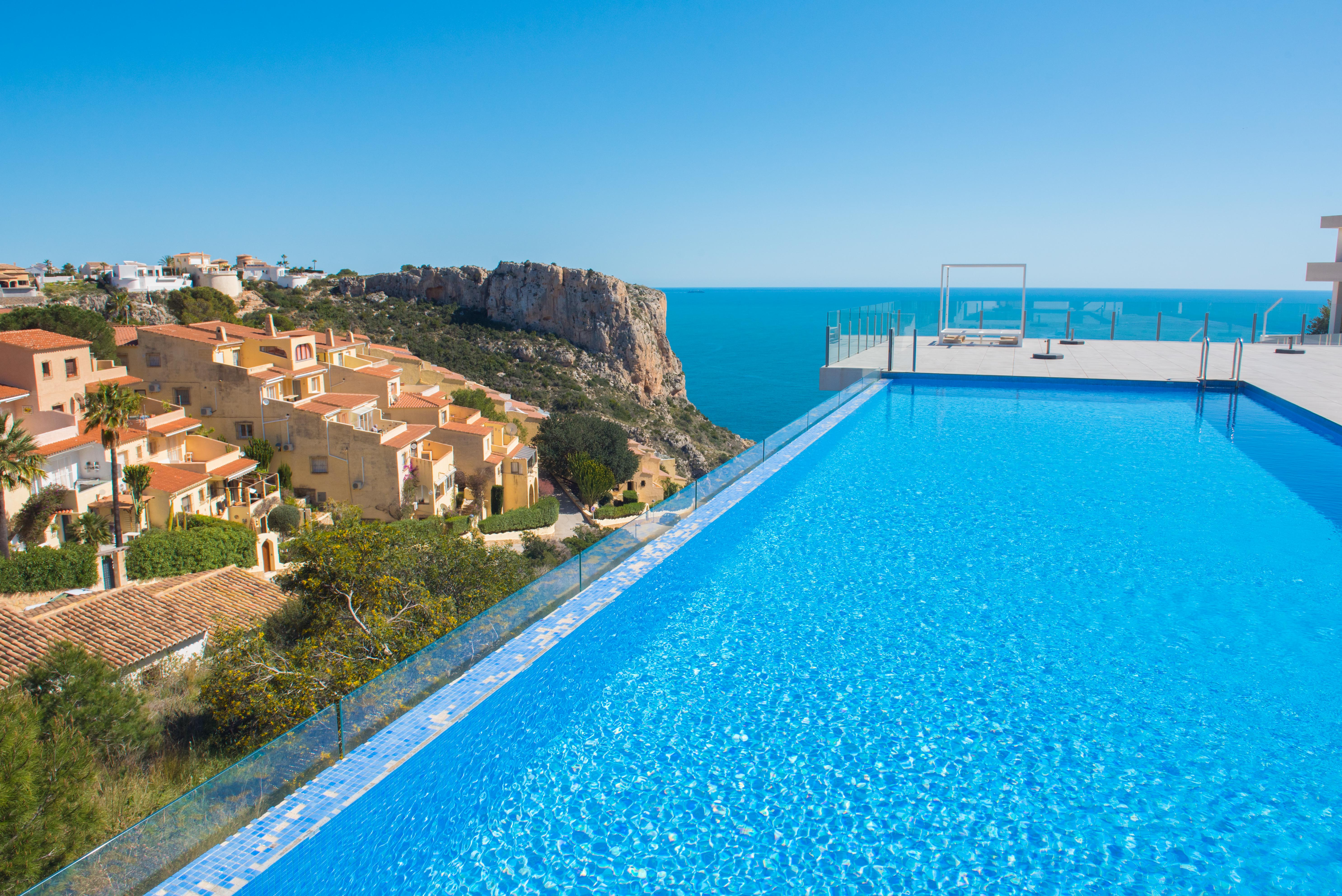 Piscina infinita con revestimiento de gresite azul y barandilla de cristal, orientada hacia el mar Mediterráneo y los acantilados de Benitachell.