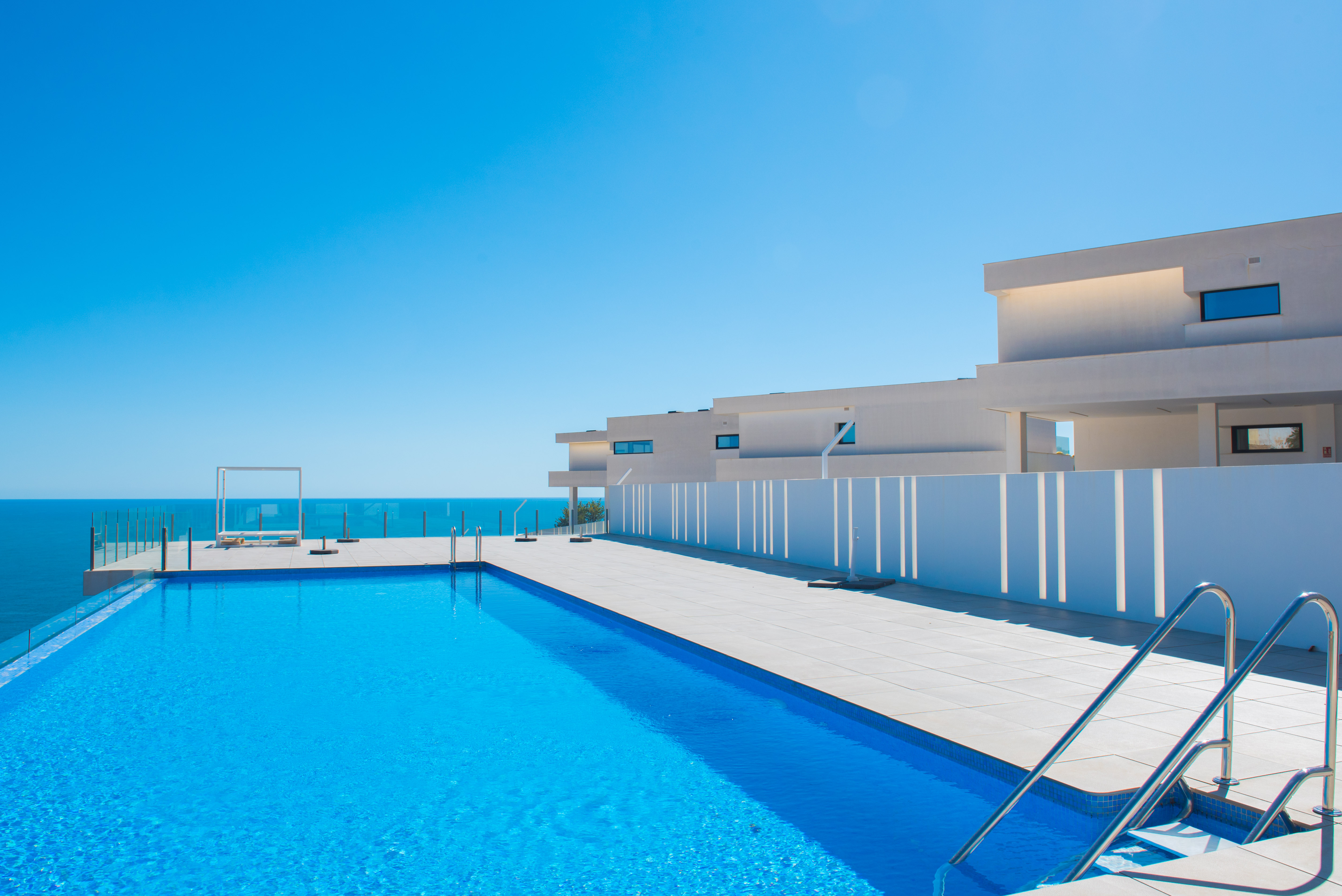 Piscina infinita con vistas al Mediterráneo, rodeada de una amplia terraza de gres y arquitectura minimalista blanca con barandillas de cristal.