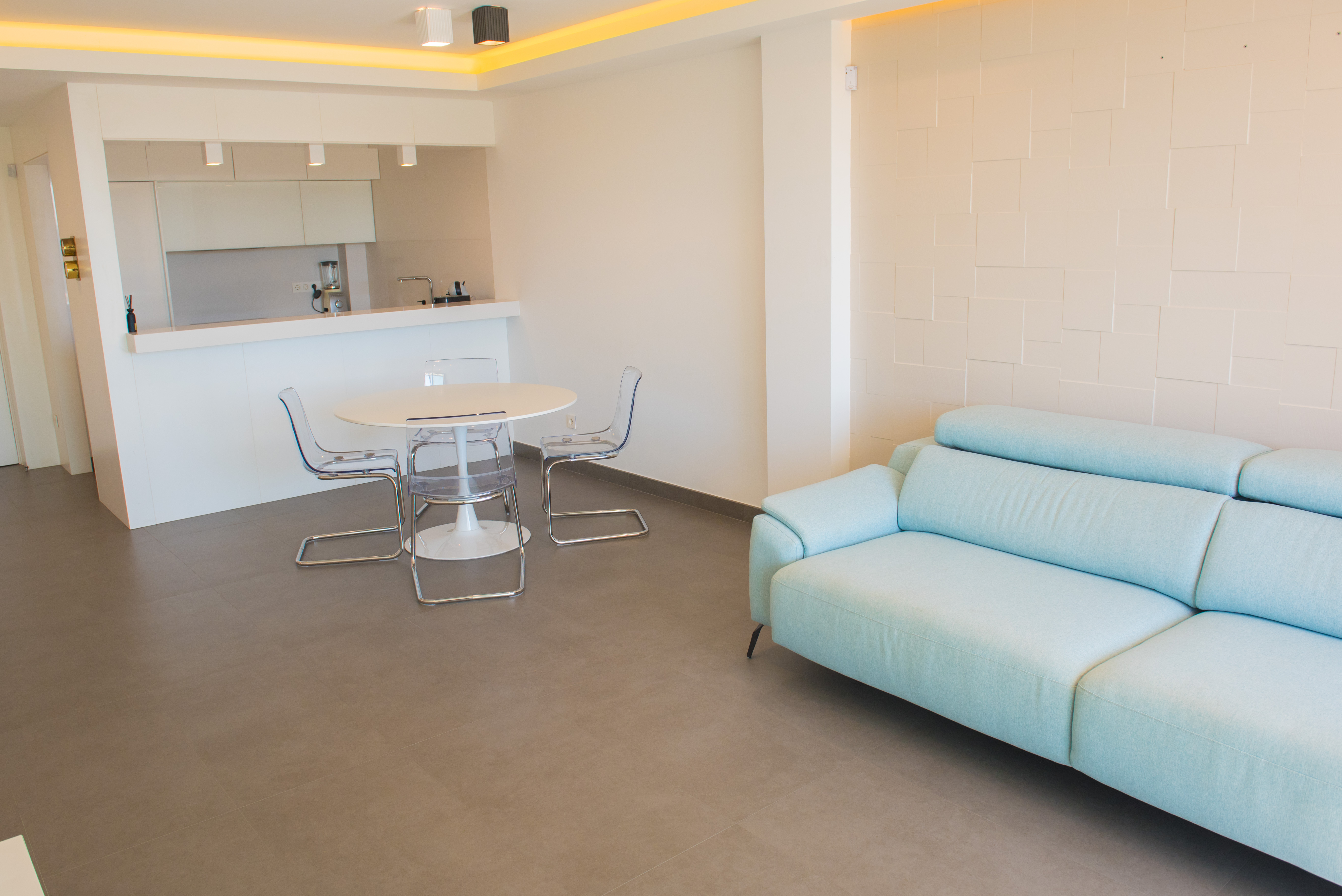 Salón-comedor diáfano con sofá azul claro, mesa redonda y sillas de metacrilato. Destaca la pared con relieve geométrico y la iluminación indirecta en el techo.