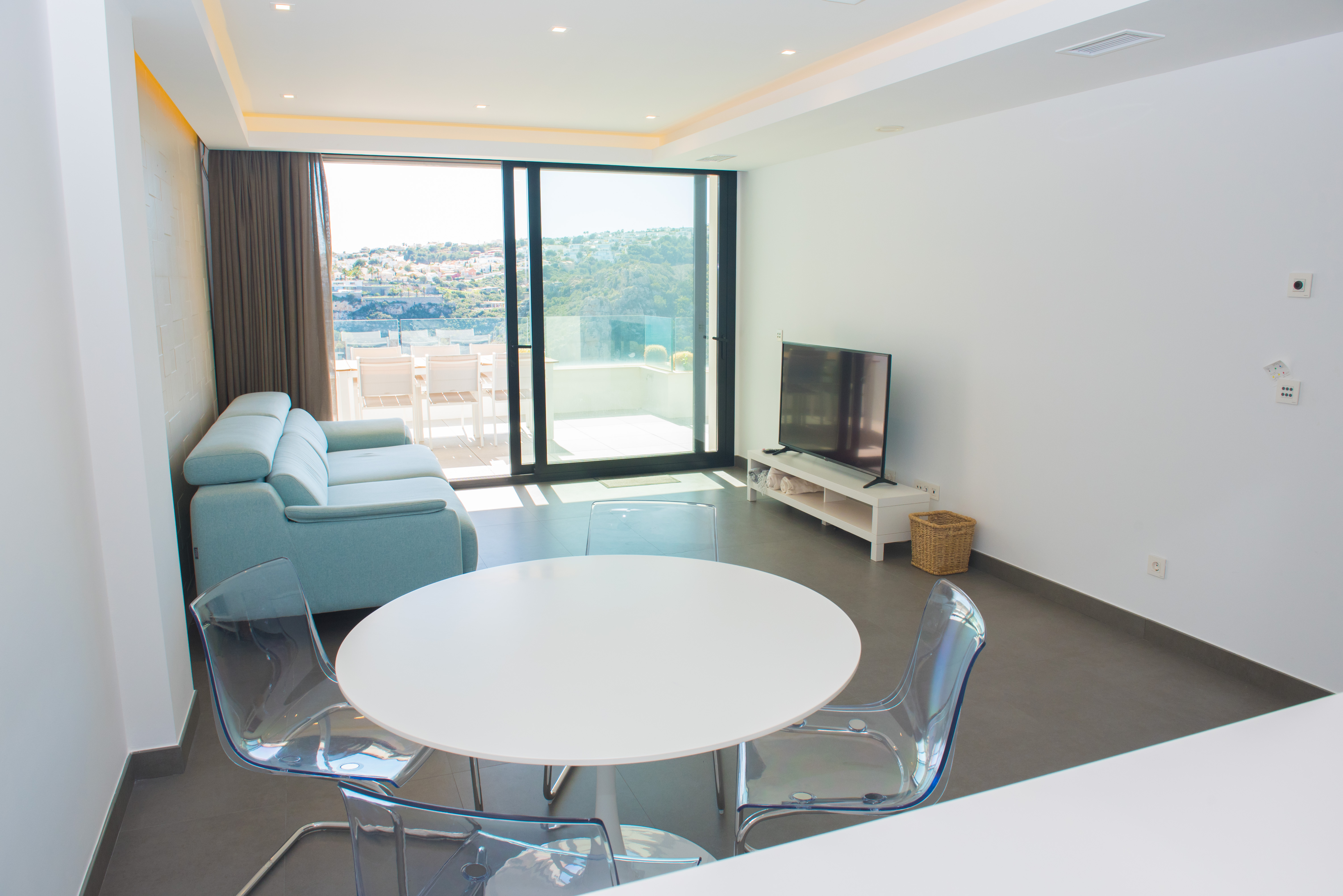 Salón-comedor minimalista con mesa circular blanca, sillas acrílicas y sofá azul claro, integrado con una terraza exterior de vistas despejadas mediante grandes ventanales.