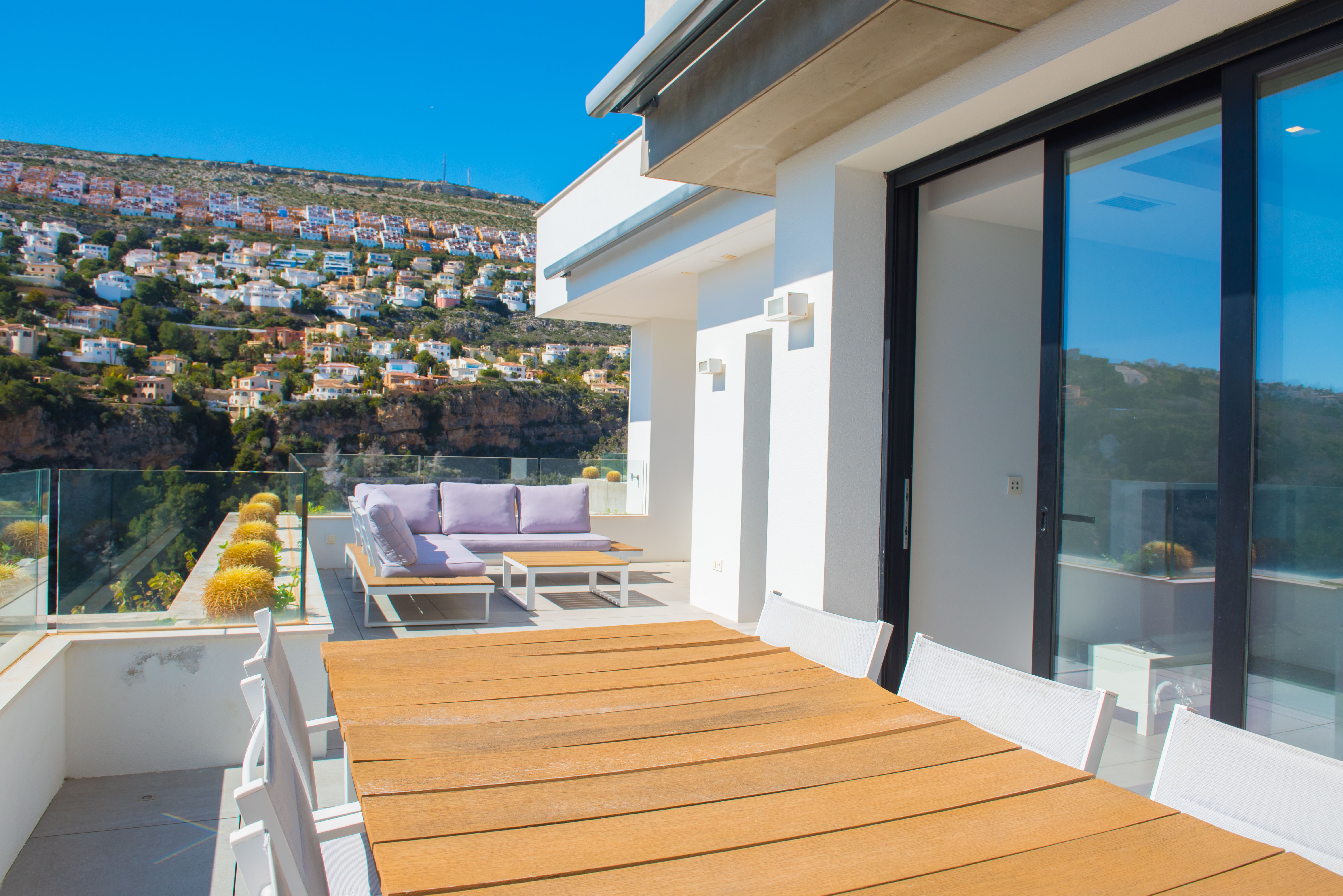 Terraza exterior con mesa de comedor de madera, zona de estar con sofás lilas y barandillas de cristal con vistas a la ladera de Benitachell.