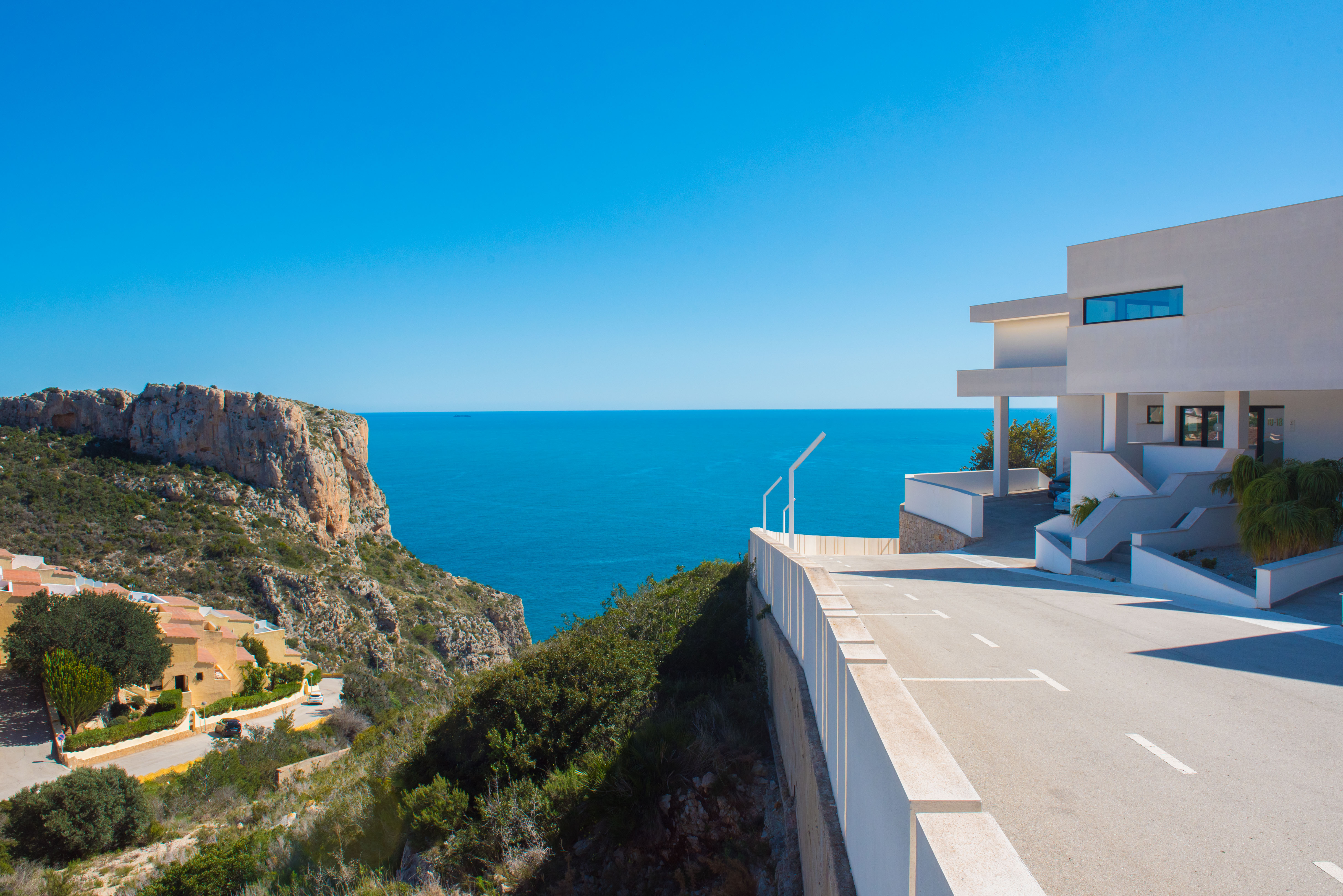 Vista exterior del complejo de arquitectura minimalista blanca, ubicado sobre un acantilado con acceso pavimentado y vistas despejadas al mar Mediterráneo.