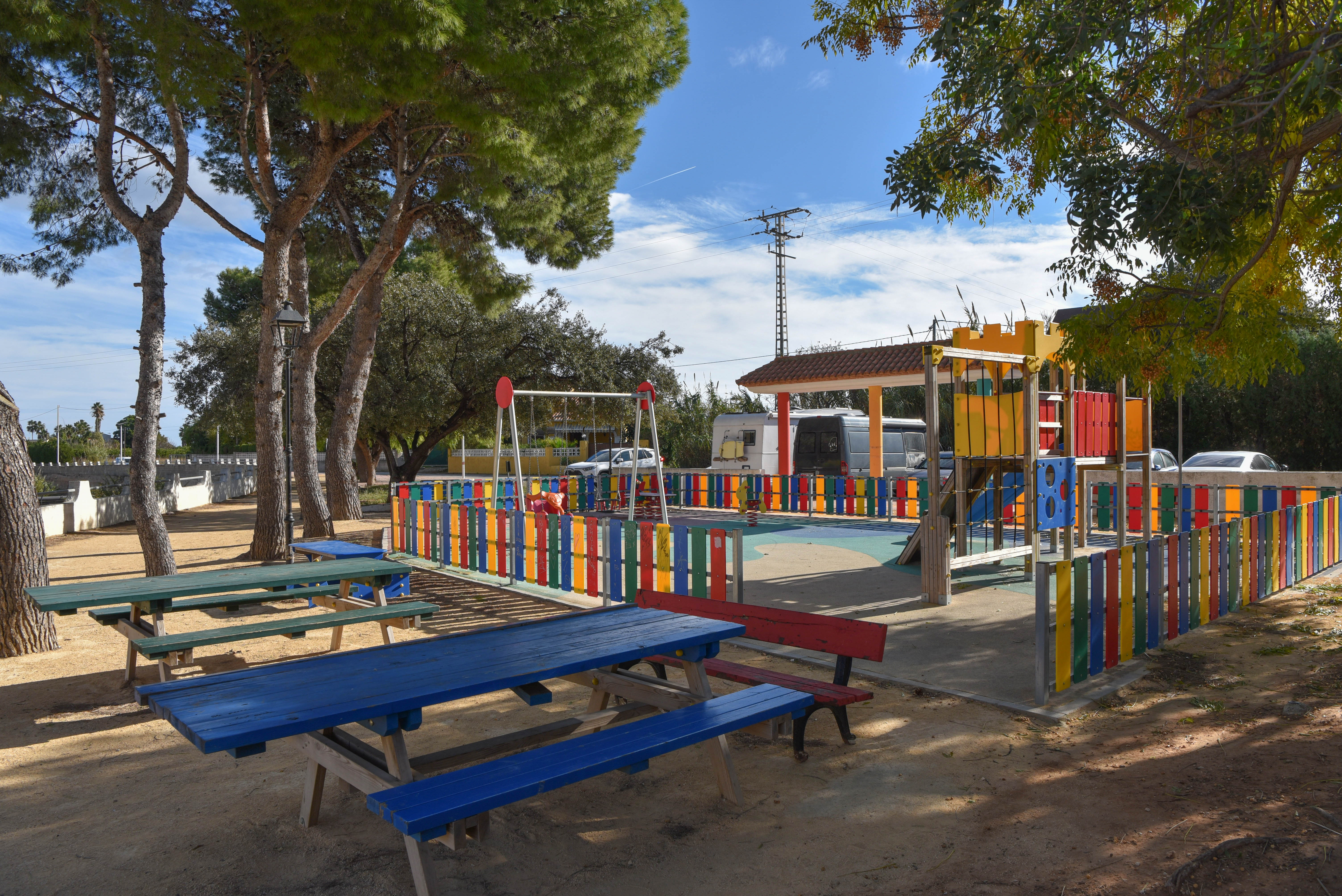 Área recreativa infantil vallada con juegos coloridos y mesas de picnic de madera bajo la sombra de pinos maduros en un entorno urbano.