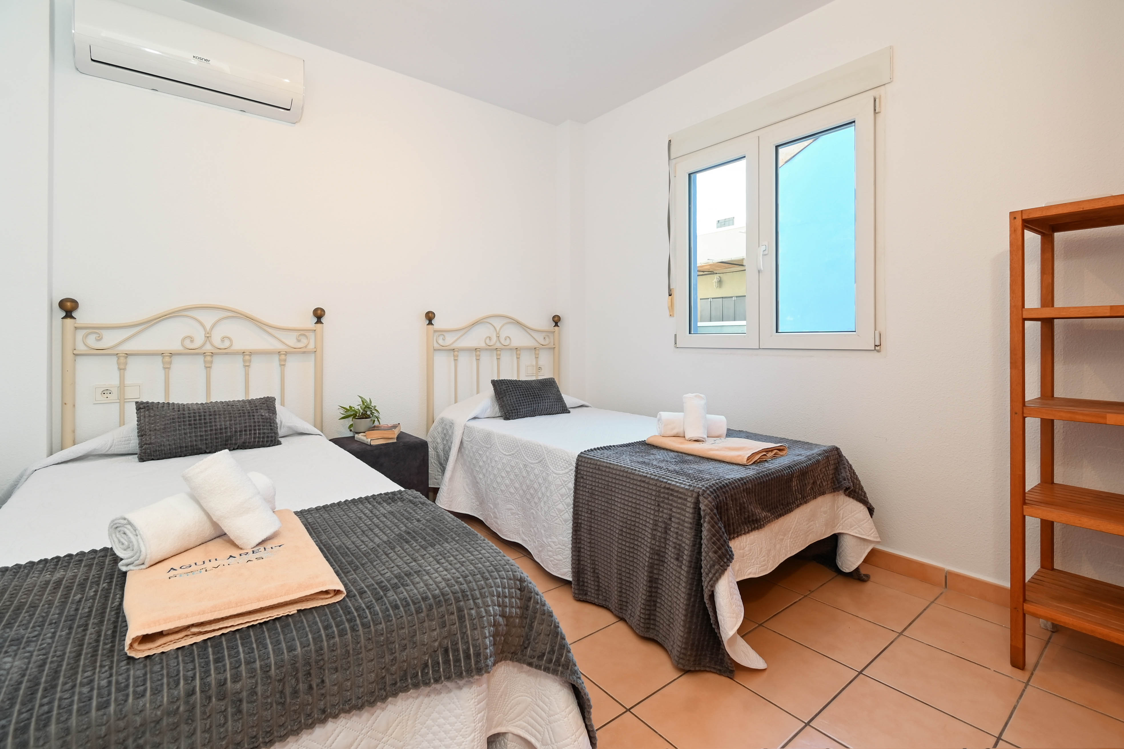 Dormitorio con dos camas individuales, cabeceros de hierro forjado, suelos de gres rústico y aire acondicionado. Incluye estantería de madera y ventana de aluminio blanco.