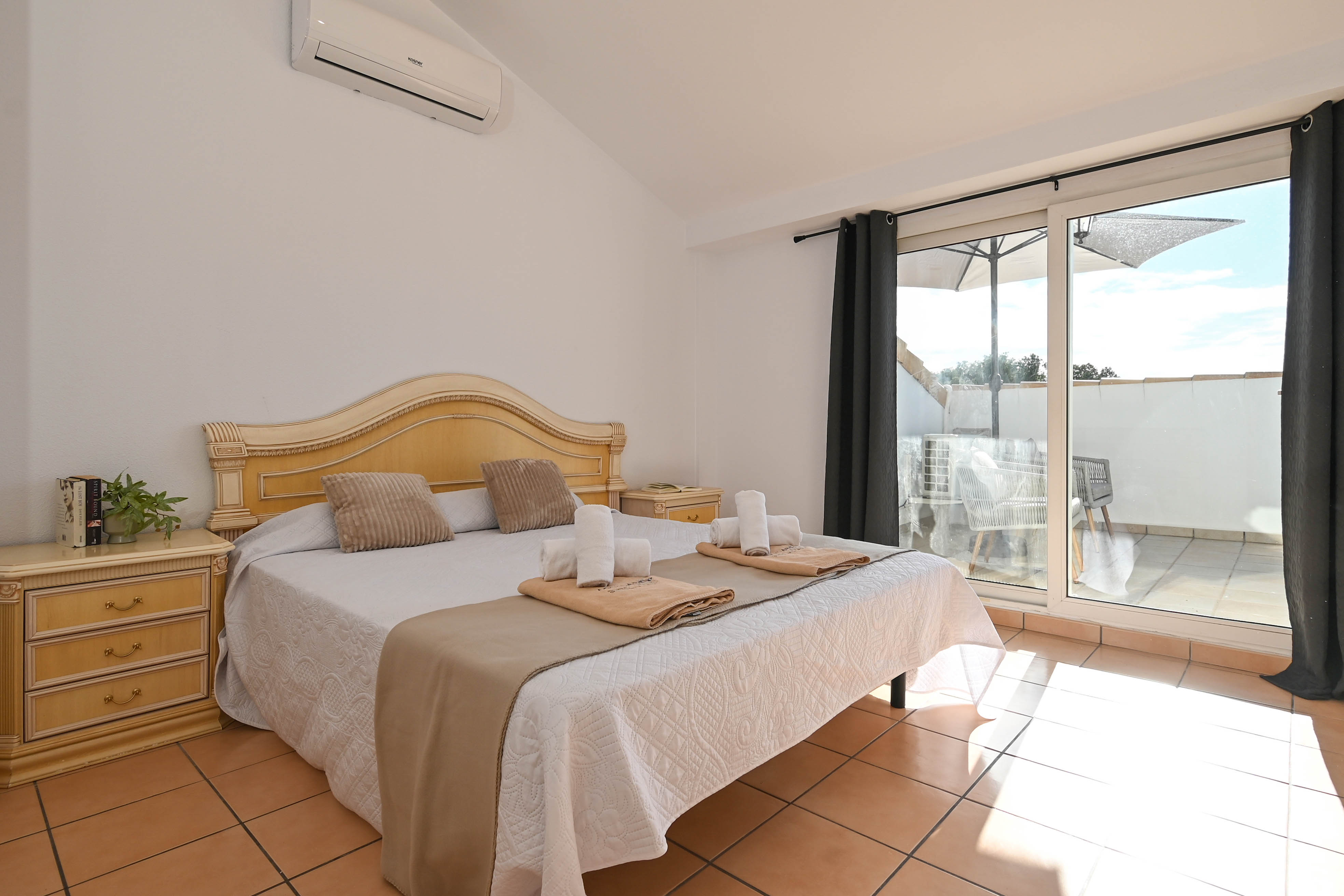 Dormitorio con techo inclinado, aire acondicionado y acceso directo a una terraza privada. Cuenta con suelos de baldosa y mobiliario de madera clásico.
