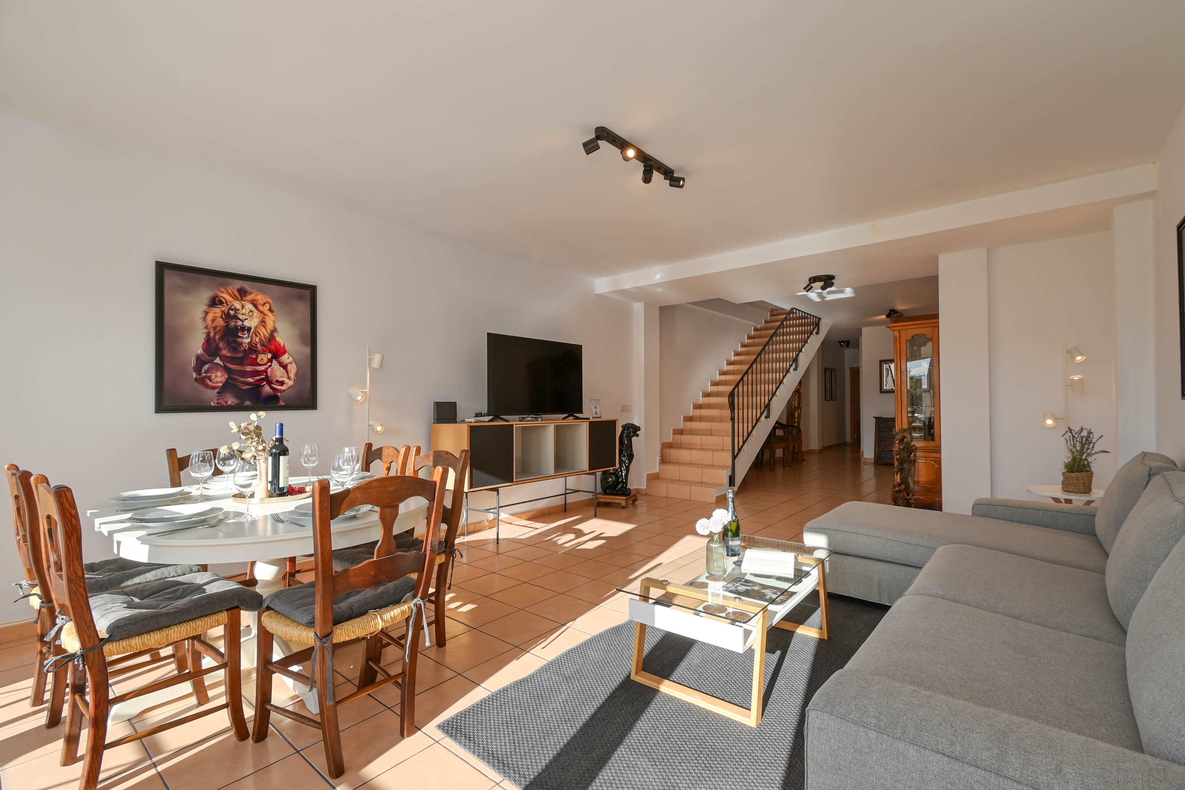 Salón-comedor de planta abierta con suelos de gres, mobiliario de madera, sofá en L y una escalera con barandilla metálica que conecta con el nivel superior.