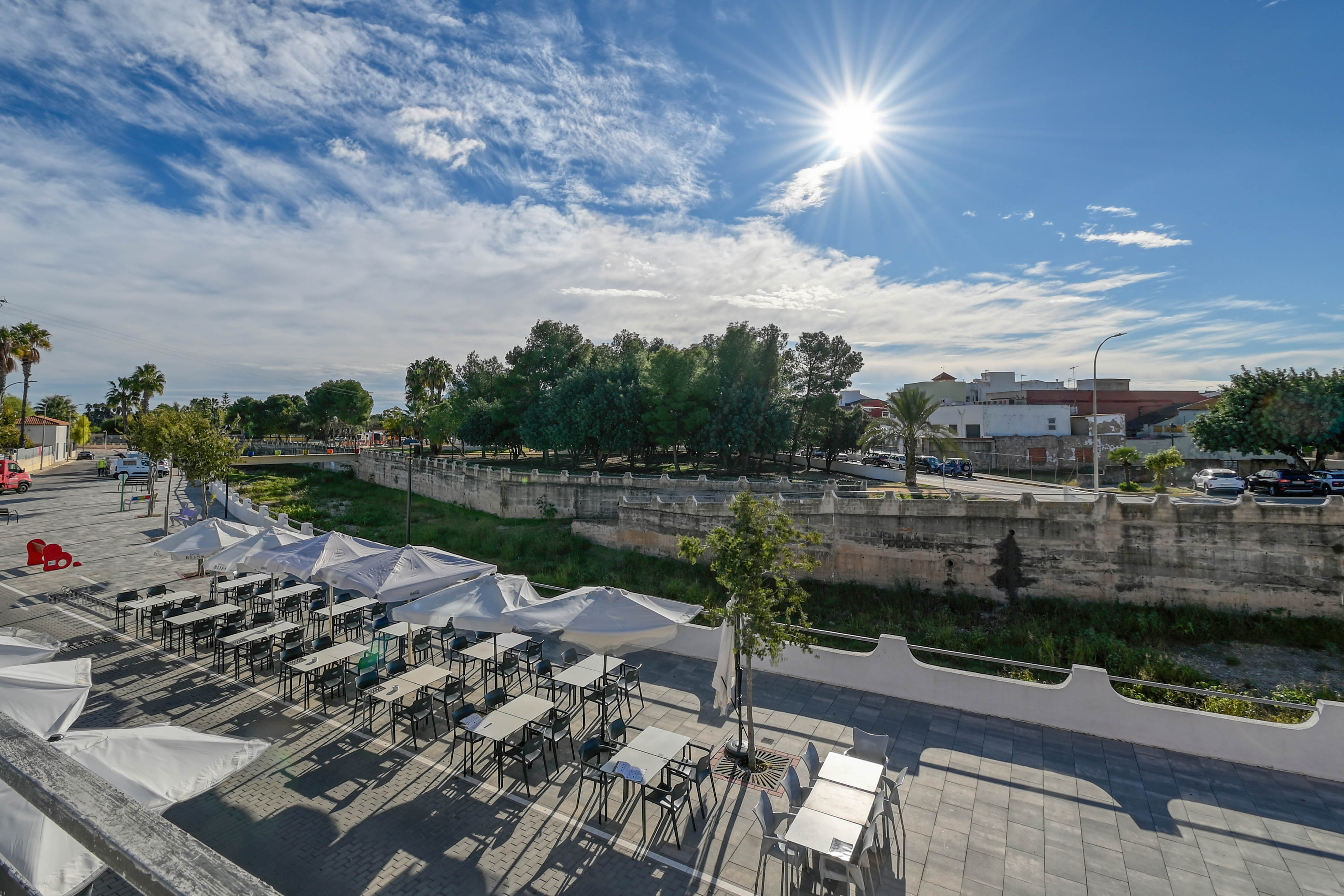 Terraza exterior con mesas, sillas y sombrillas blancas junto a un paseo urbano y zona arbolada bajo un cielo despejado y soleado.