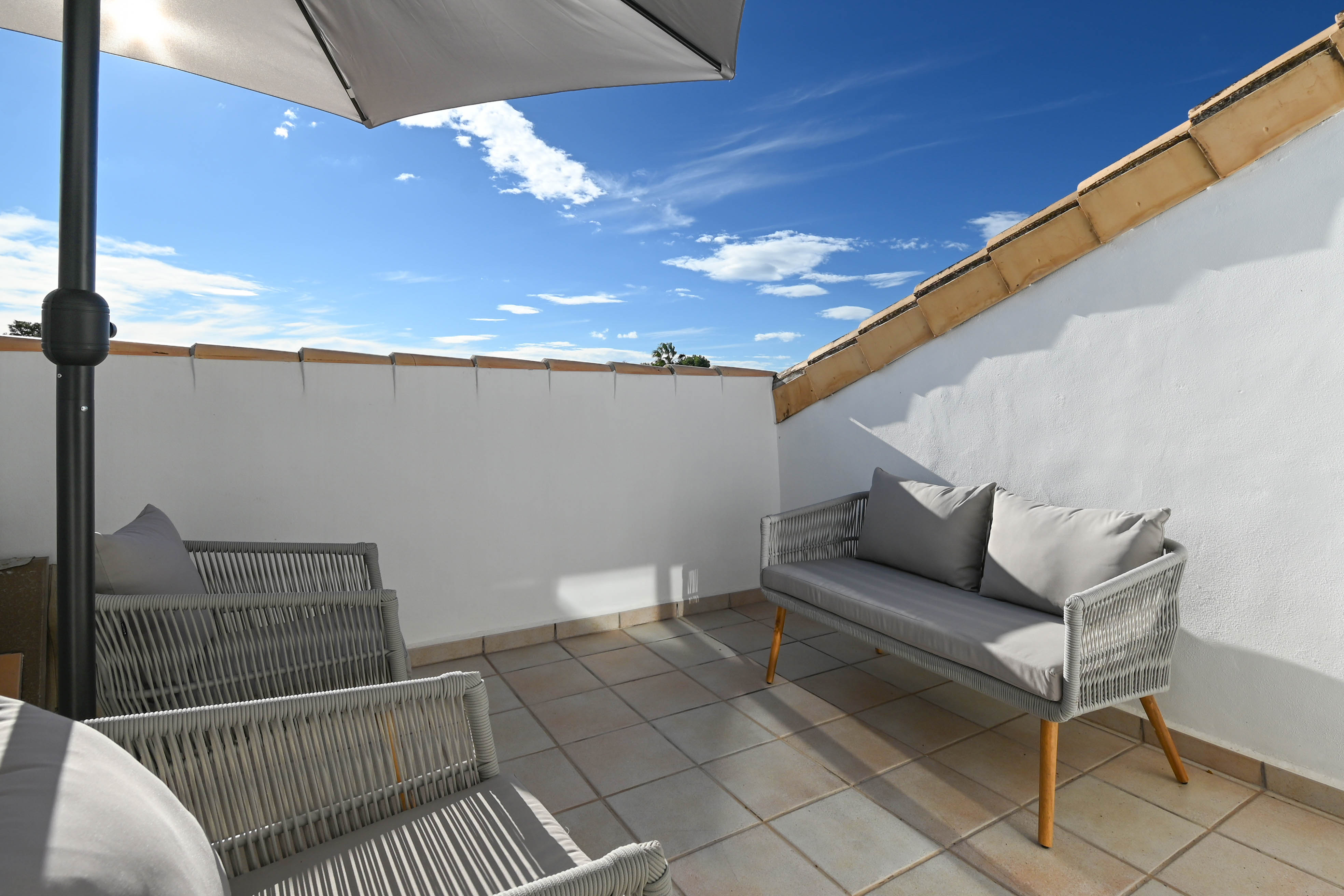 Terraza privada en la azotea con mobiliario de exterior en tonos grises, suelo cerámico y sombrilla, bajo el cielo despejado de la Costa Blanca.