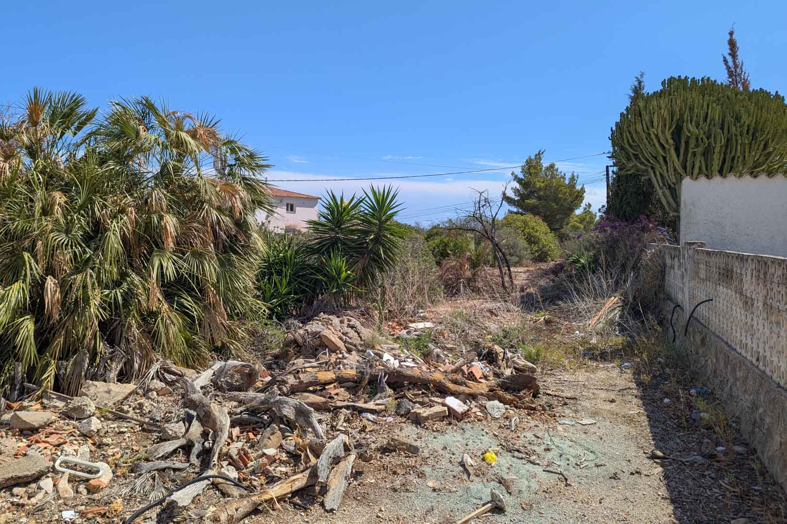 Parcela urbana con vegetación mediterránea densa, restos de madera en primer plano y un muro perimetral bajo un cielo despejado en la zona del Montgó.