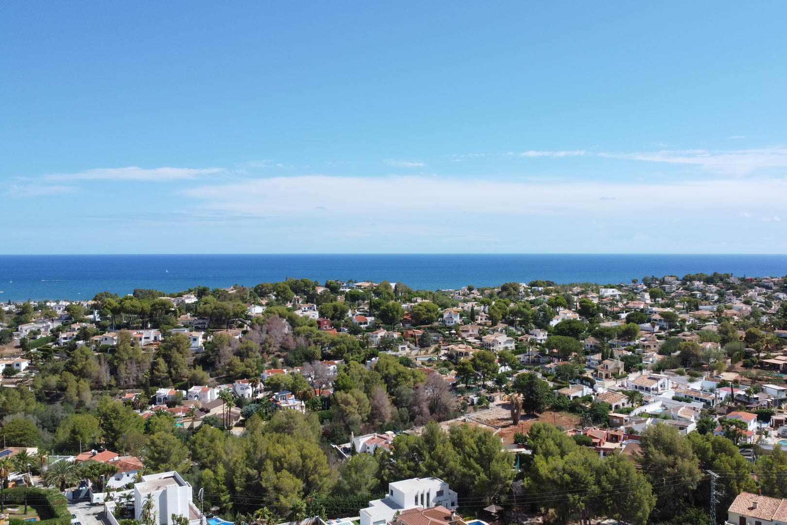Vista aérea panorámica de una zona residencial en Dénia, con viviendas unifamiliares rodeadas de pinos mediterráneos y el horizonte del mar al fondo.