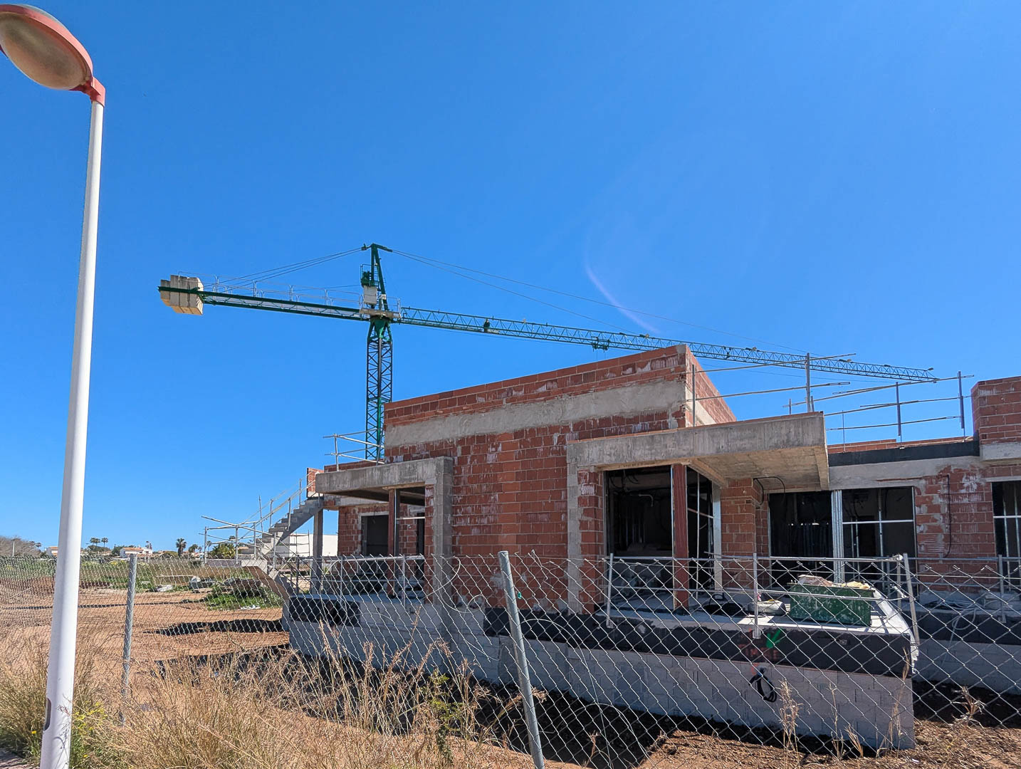Vista de la fase de construcción actual con estructura de ladrillo y hormigón, destacando la grúa de obra y el vallado perimetral bajo un cielo despejado.