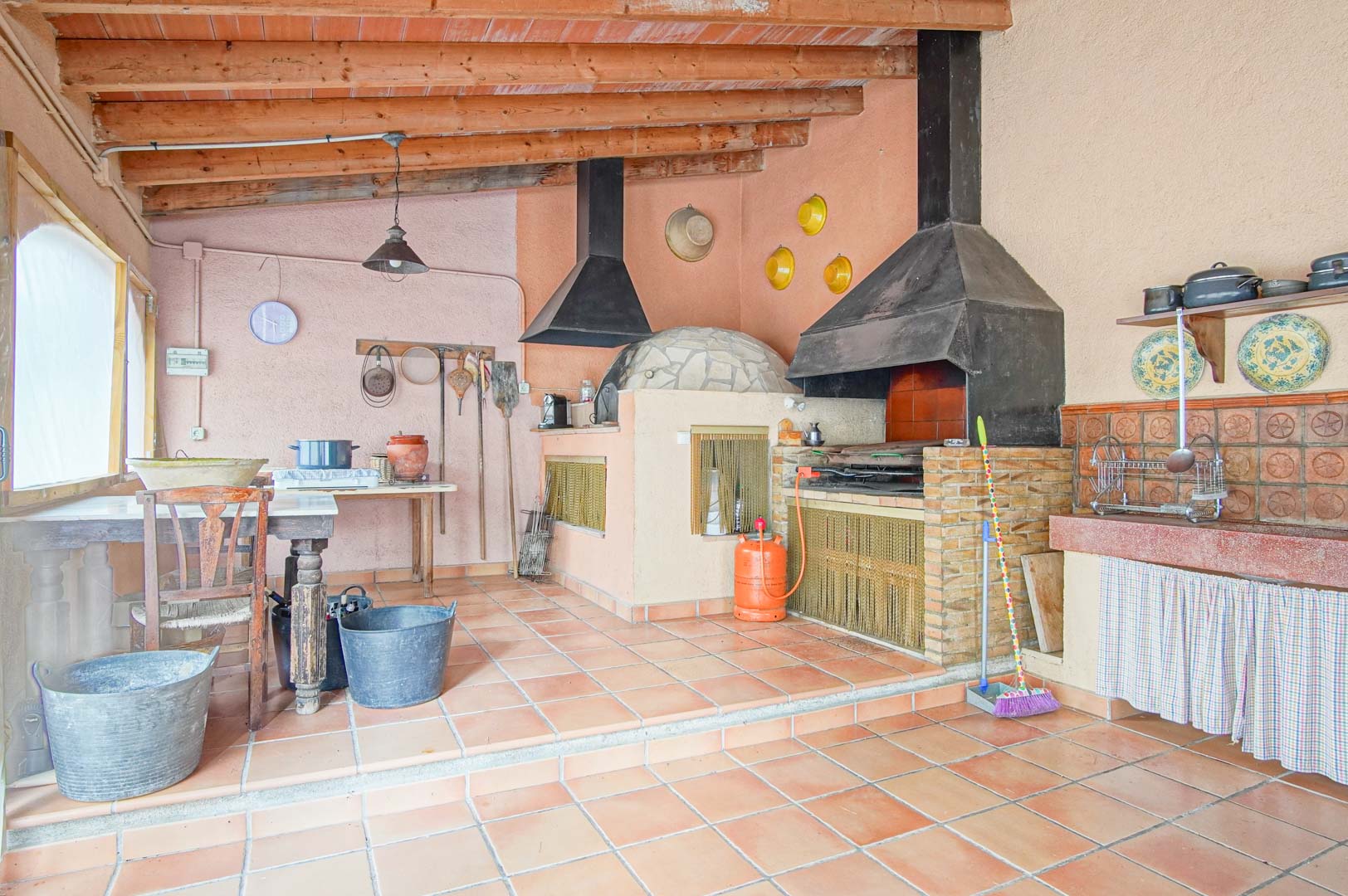 Cocina rústica exterior con horno de leña tradicional, barbacoa de obra y techos de vigas de madera sobre suelos de barro cocido.