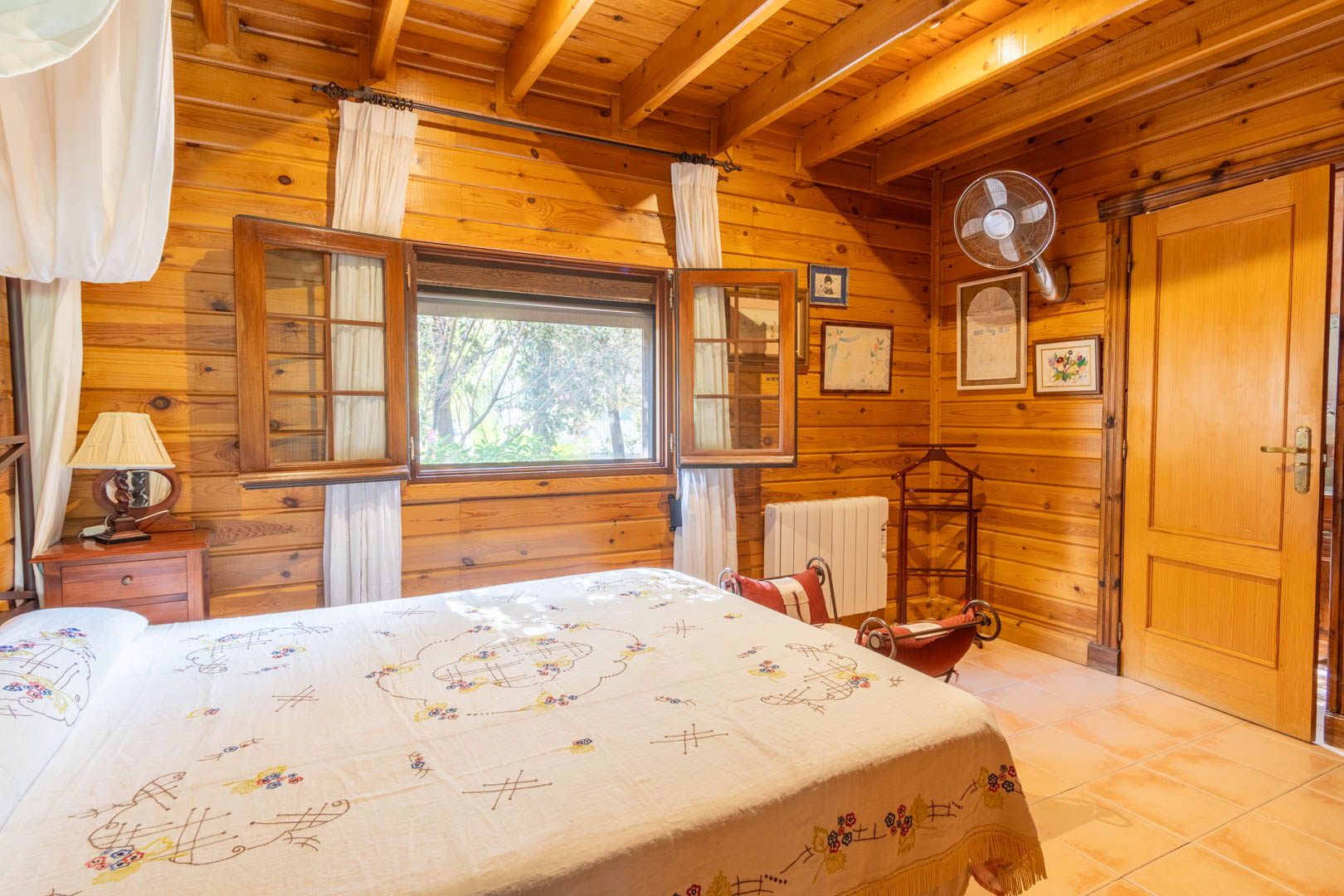 Dormitorio rústico revestido íntegramente en madera de pino finlandés, con techos de vigas vistas, suelos cerámicos y ventana con contraventanas de madera que ofrece luz natural.