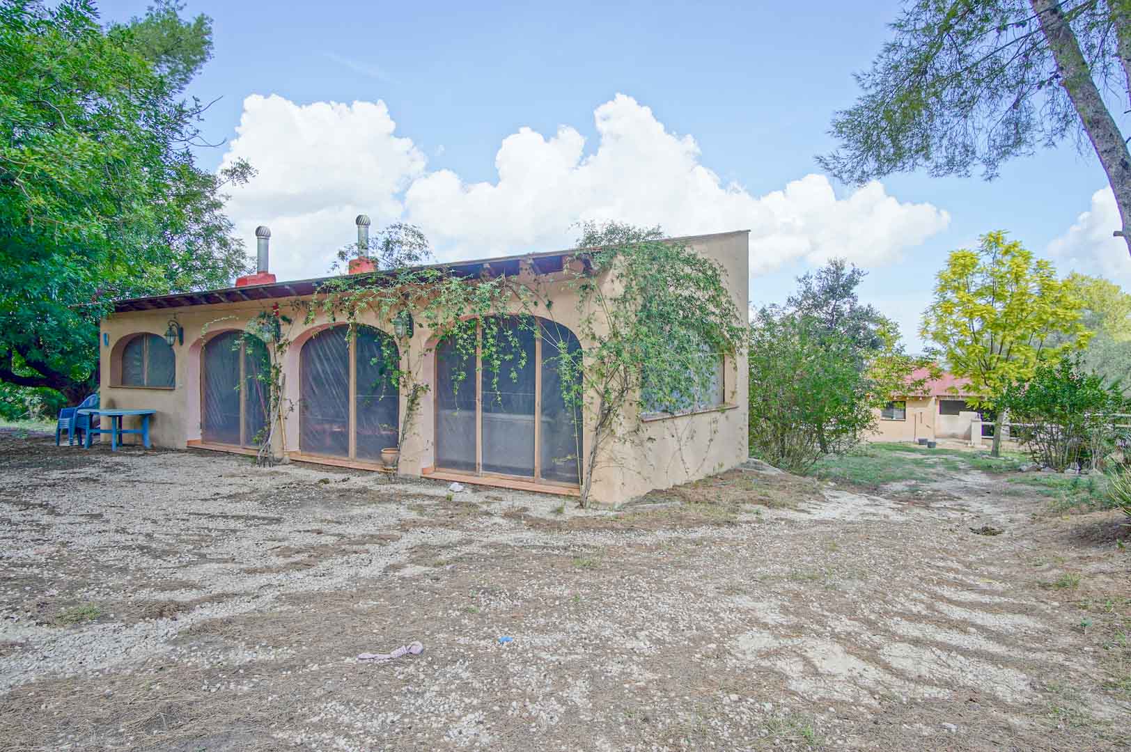 Edificación exterior con arcos protegidos y vigas de madera vista, rodeada de pinos y vegetación mediterránea en una parcela natural de Orba.