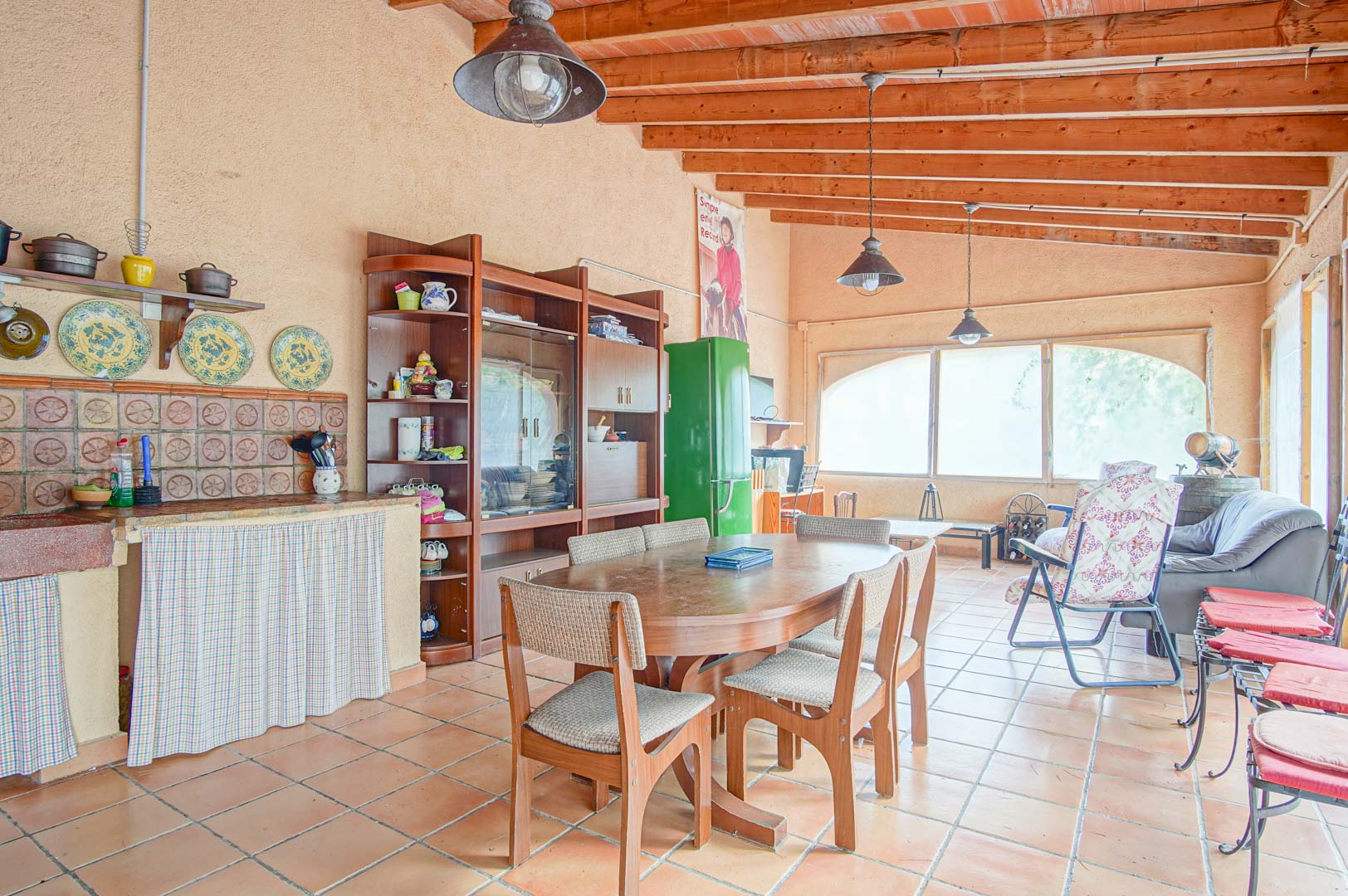 Espaciosa zona de reuniones rústica con techos de vigas de madera, suelos de barro, cocina integrada y comedor iluminado por amplios ventanales.