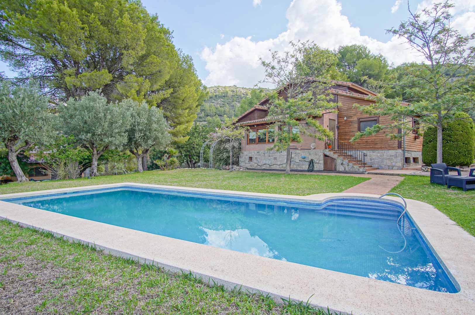 Exterior de la villa con piscina rectangular rodeada de césped. Se observa la fachada de madera de pino y piedra natural, integrada en un entorno de pinos y montañas.