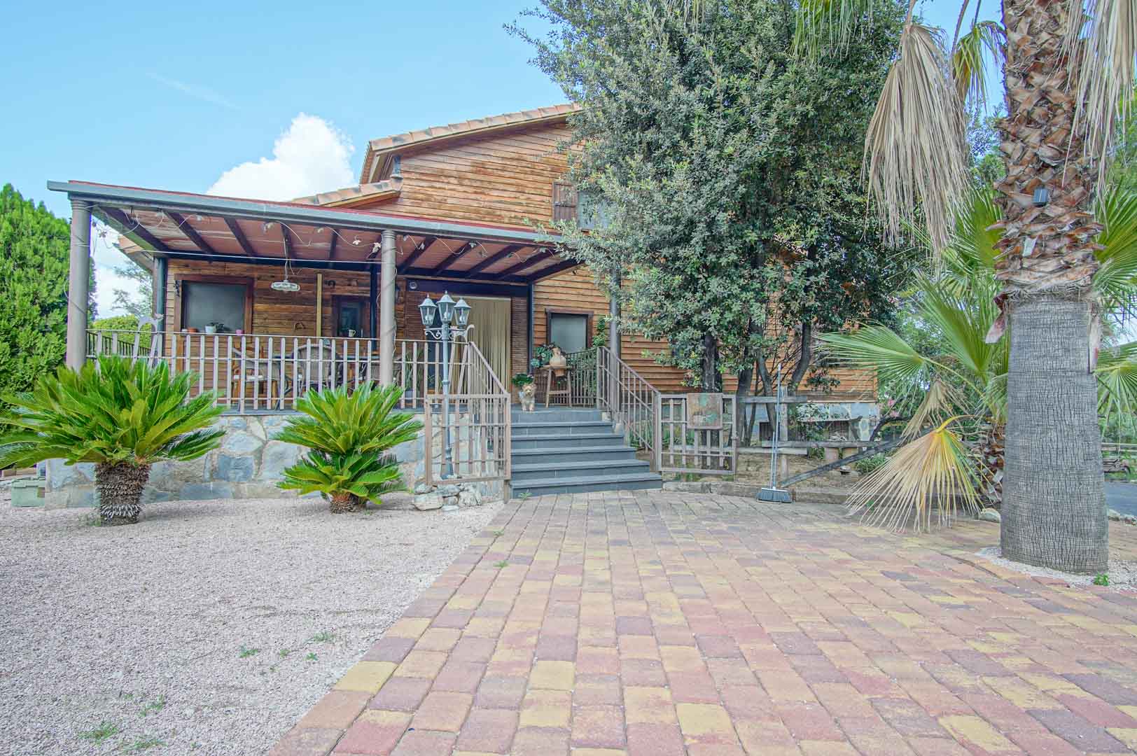 Fachada de madera de pino finlandés con porche elevado, base de piedra natural y escalinata de acceso rodeada de vegetación mediterránea y palmeras.