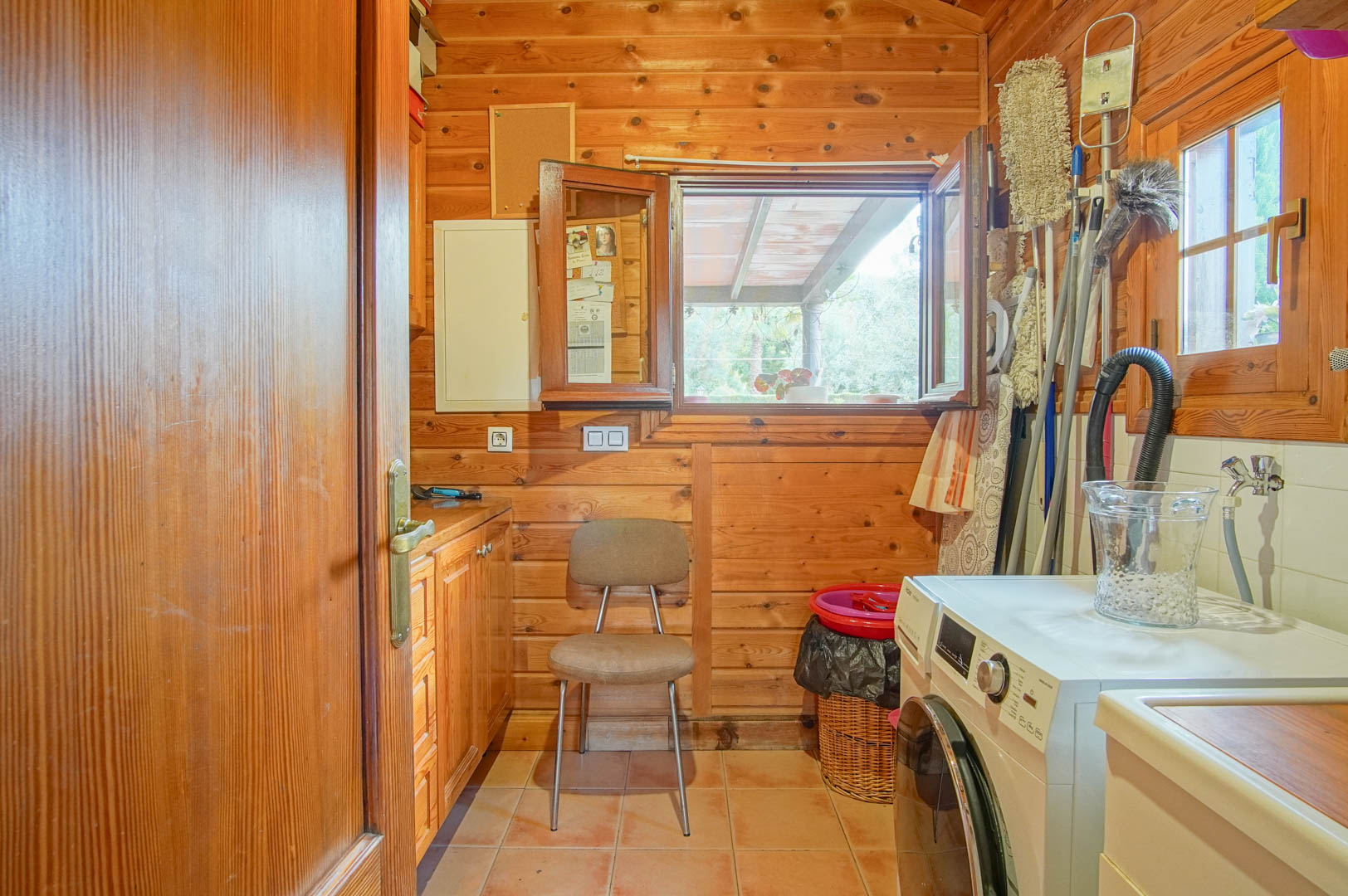 Lavadero funcional revestido en madera de pino finlandés, equipado con lavadora, zona de almacenaje y ventana con vistas al jardín.