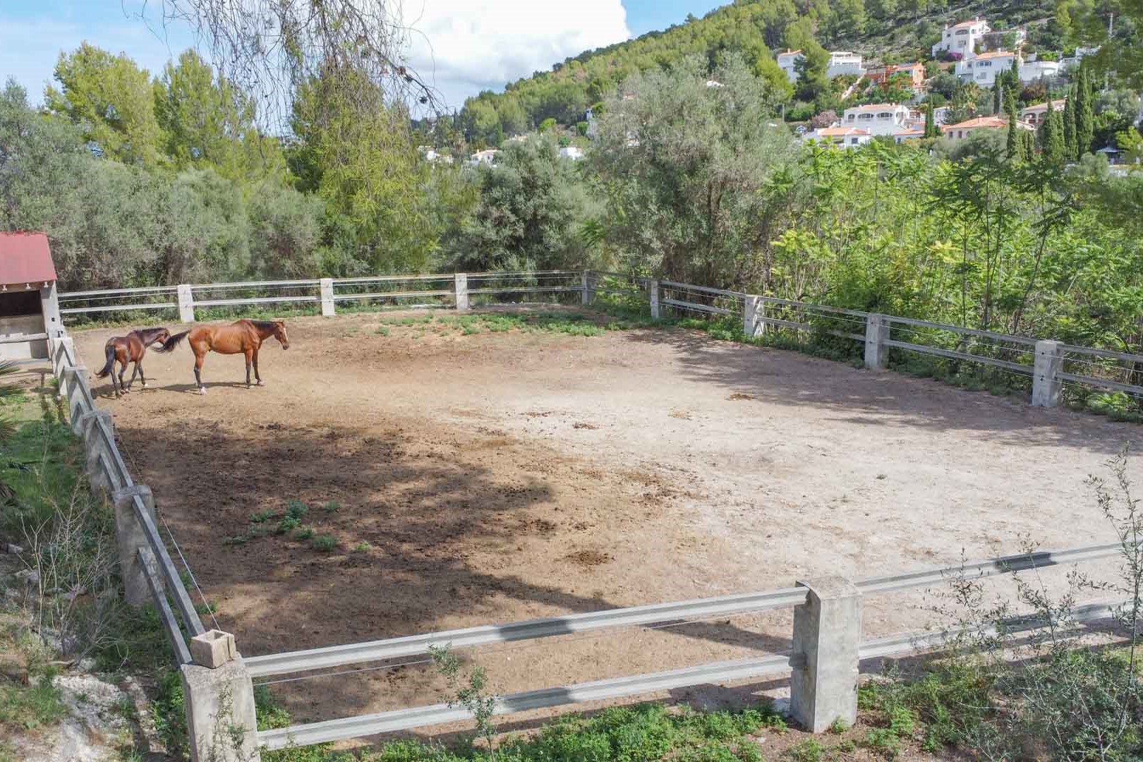Pista ecuestre vallada con postes de piedra, situada en una parcela de 18.000 m² rodeada de vegetación mediterránea y vistas a la montaña.