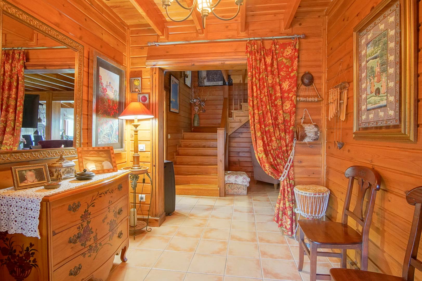 Recibidor rústico con paredes de pino finlandés, suelos cerámicos y una escalera de madera que conecta las plantas. Destaca la decoración cálida y el mobiliario artesanal.