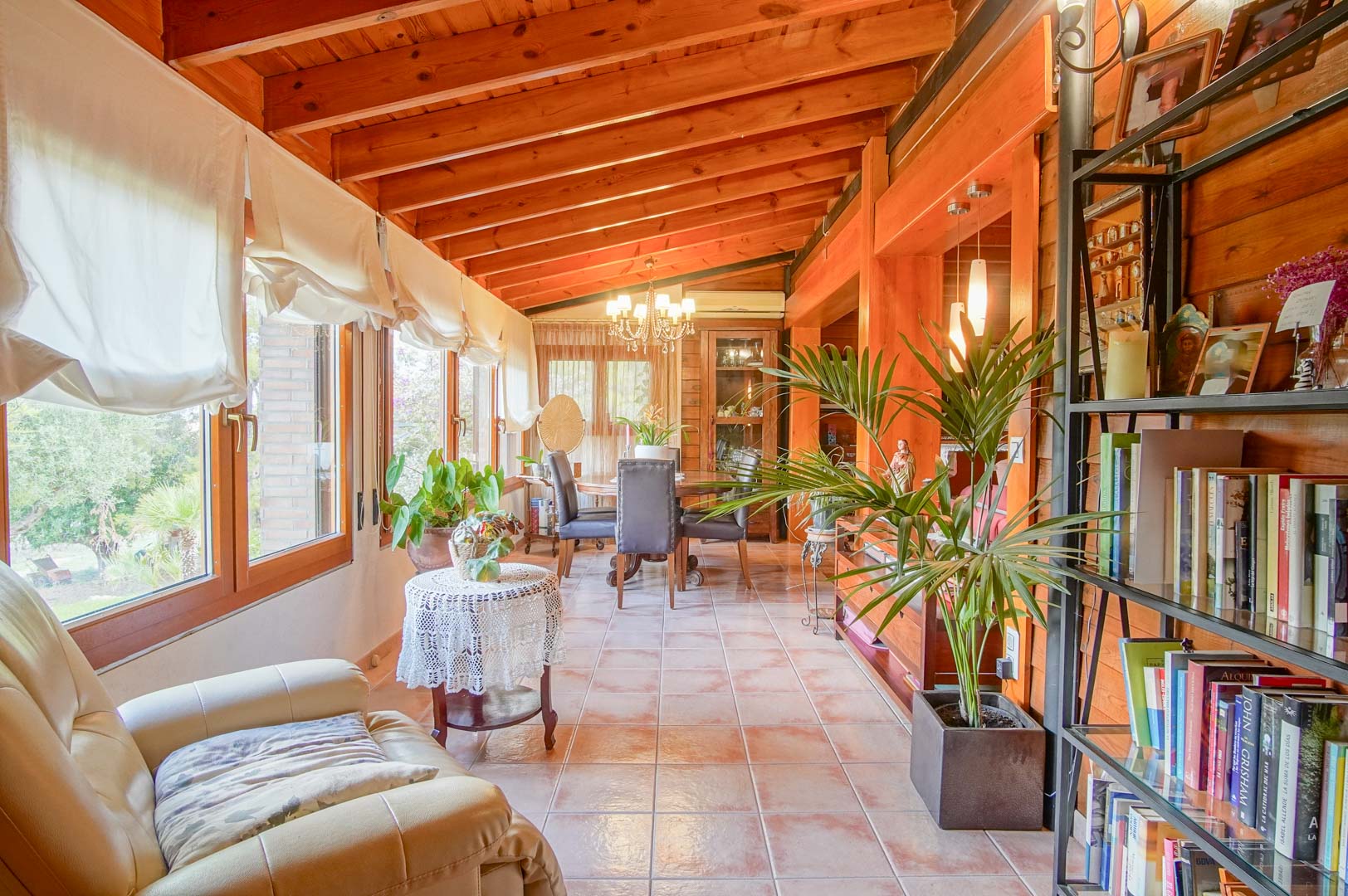 Salón acristalado con techos de vigas de pino finlandés, suelos cerámicos y amplios ventanales que ofrecen vistas al jardín mediterráneo y abundante luz natural.