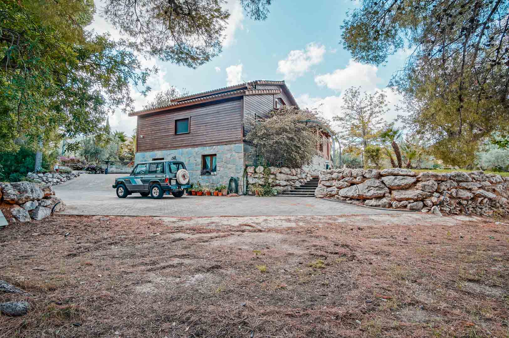Vista exterior de la villa mostrando la fachada de piedra natural y madera de pino finlandés, con amplio acceso pavimentado y muros de contención de piedra.