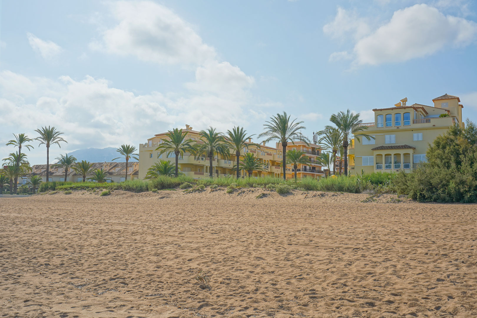 Amplia playa de arena frente a edificios residenciales de estilo mediterráneo con palmeras y vistas a la montaña bajo un cielo despejado.