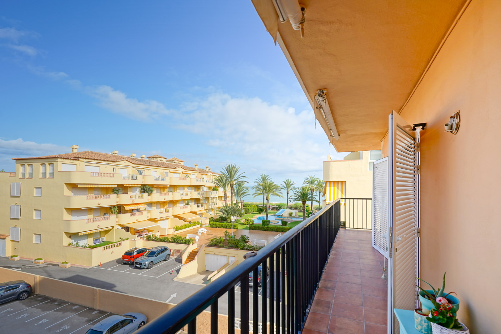Balcón con suelos de gres y barandilla metálica con vistas a la piscina comunitaria, zonas ajardinadas y el mar Mediterráneo al fondo.