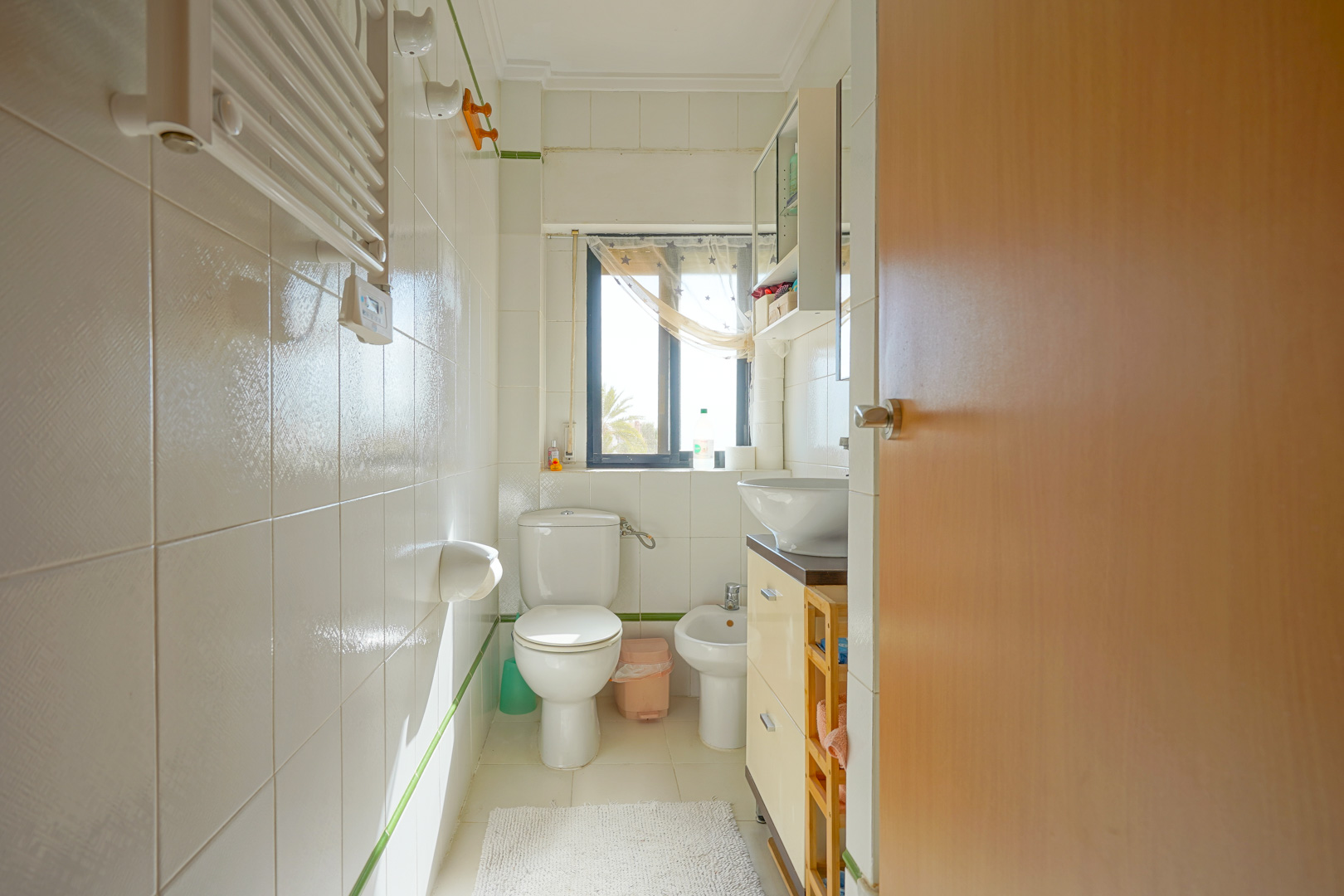 Baño funcional con azulejos blancos, bidé, inodoro y lavabo sobre encimera. Una ventana exterior aporta luz natural y vistas despejadas al entorno.