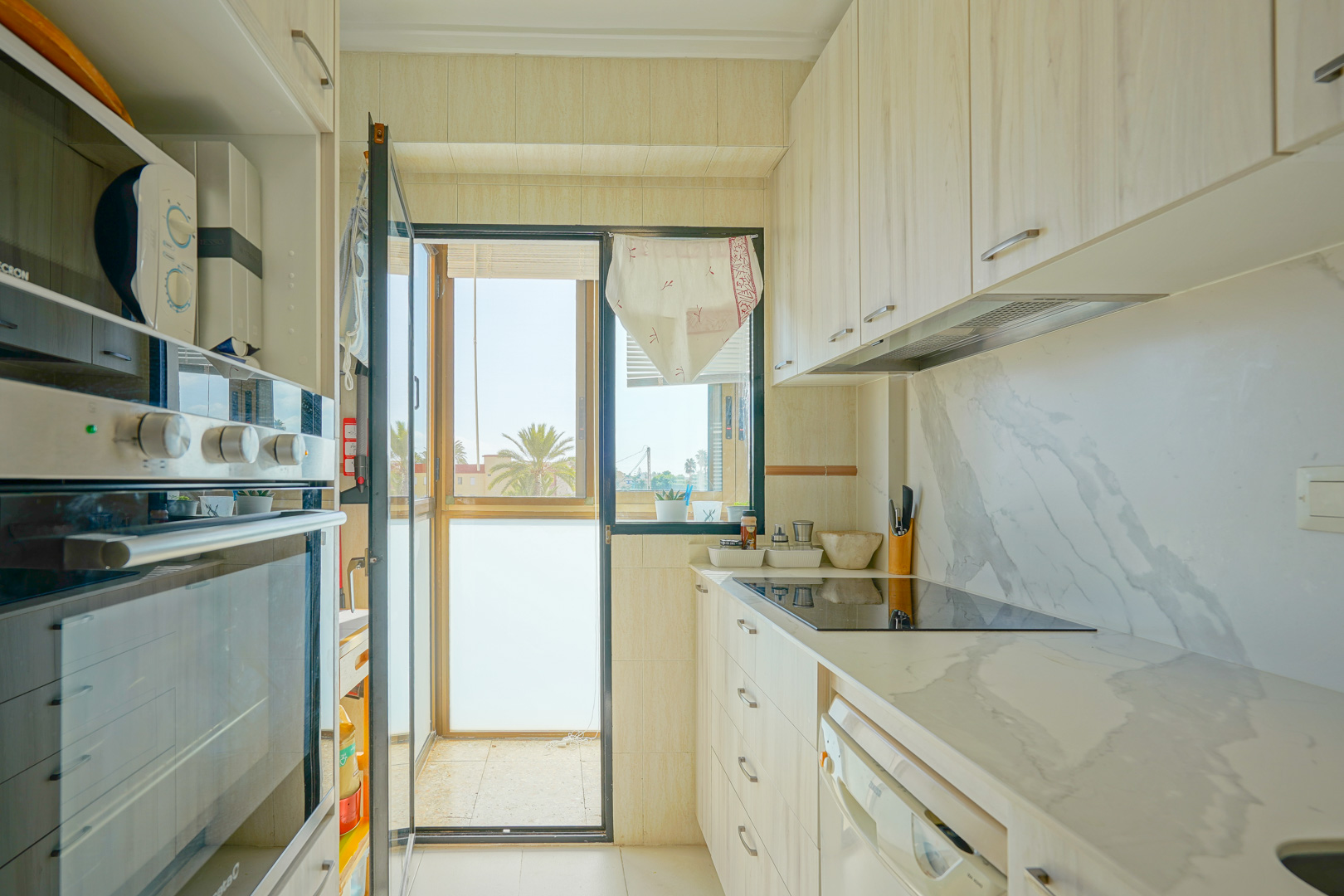 Cocina funcional con armarios de madera clara, encimera de efecto mármol y acceso directo a un balcón con vistas a palmeras.