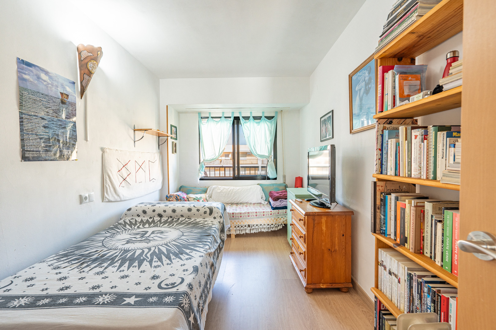 Dormitorio con cama individual, estantería de madera y cómoda. Incluye suelo laminado y una ventana con cortinas azul claro que aporta luz natural.