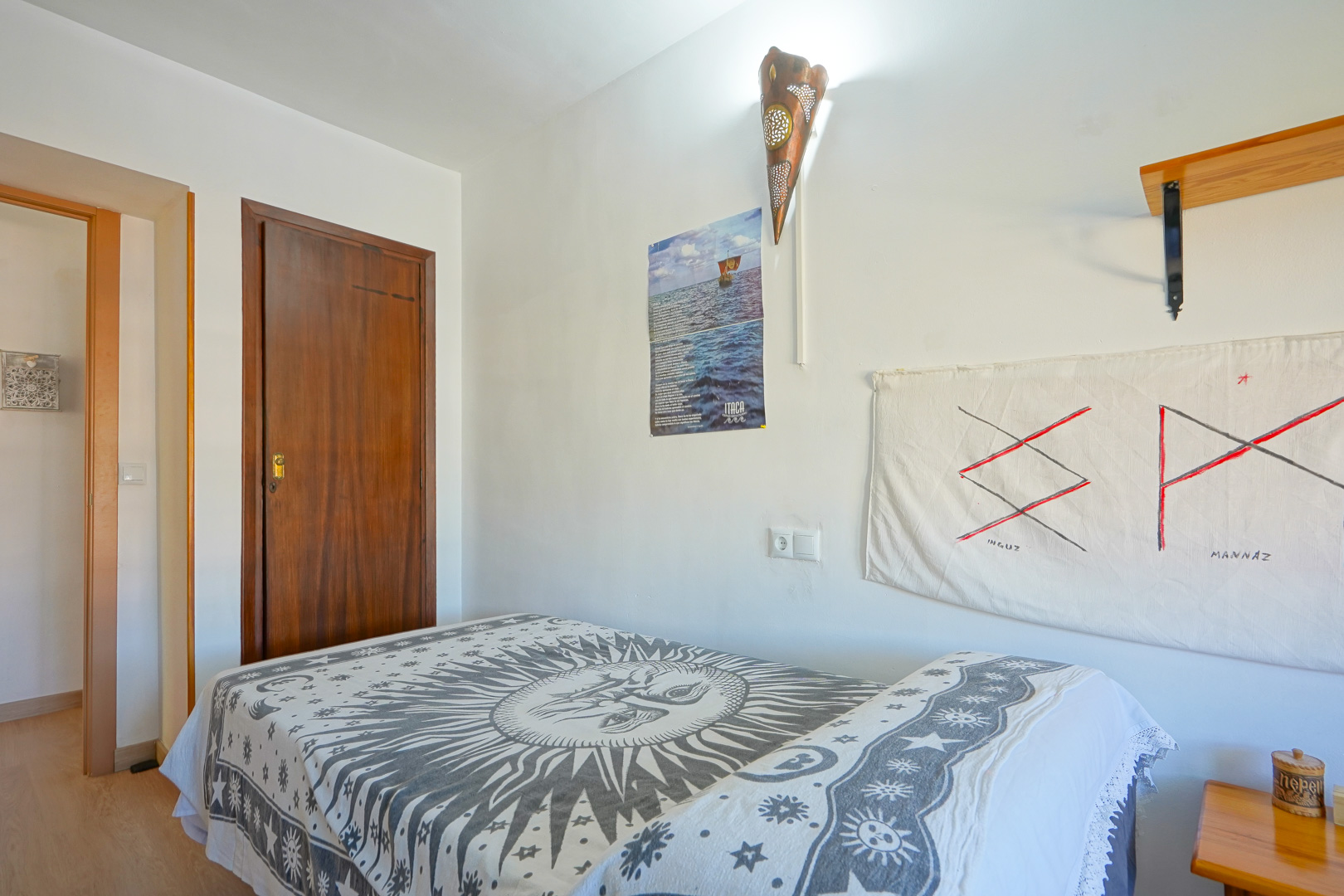 Dormitorio con cama individual y colcha de motivo solar, paredes blancas con un aplique decorativo de cobre y una puerta de madera oscura maciza.