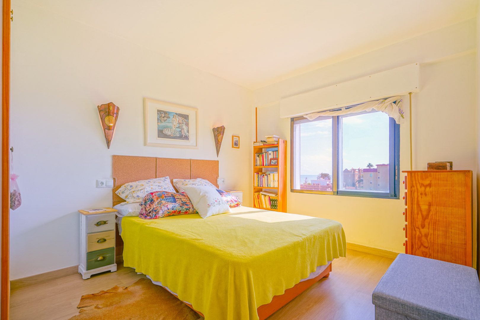 Dormitorio luminoso con cabecero de corcho, colcha amarilla y mobiliario de madera. El ventanal aporta luz natural y vistas al entorno urbano.