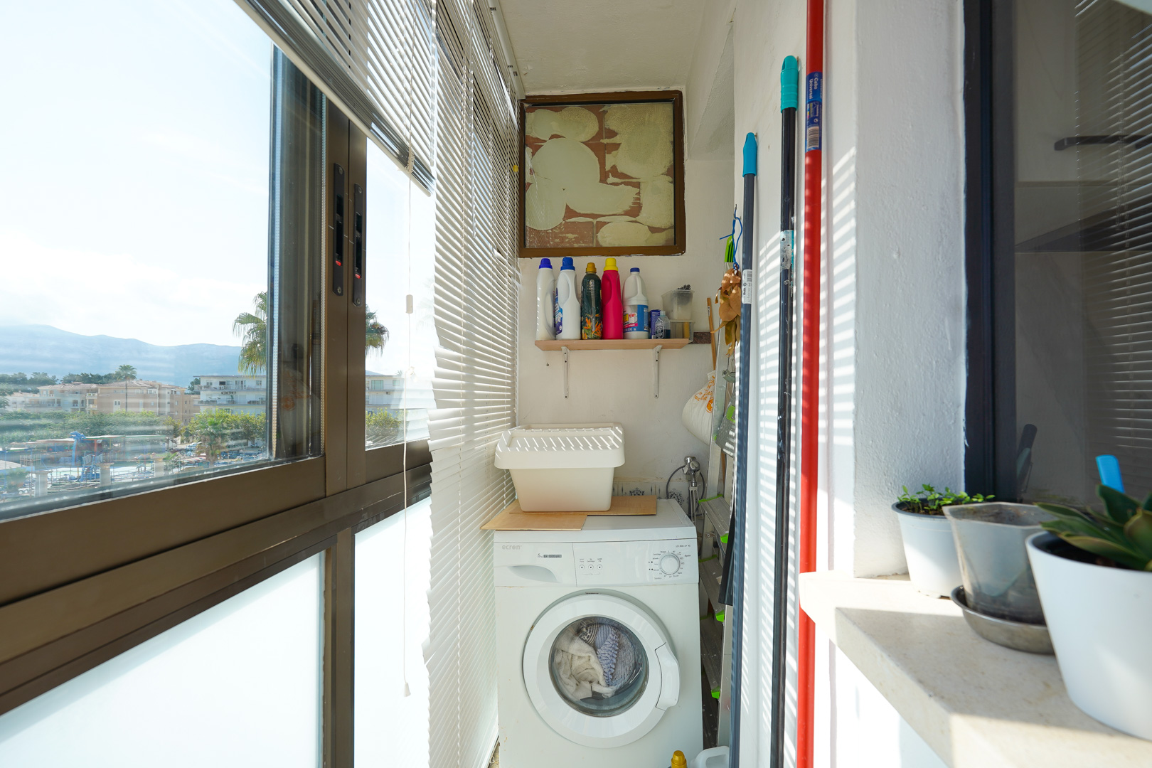 Galería cerrada con zona de lavandería que incluye lavadora de carga frontal, estantería para productos de limpieza y amplios ventanales con persianas.
