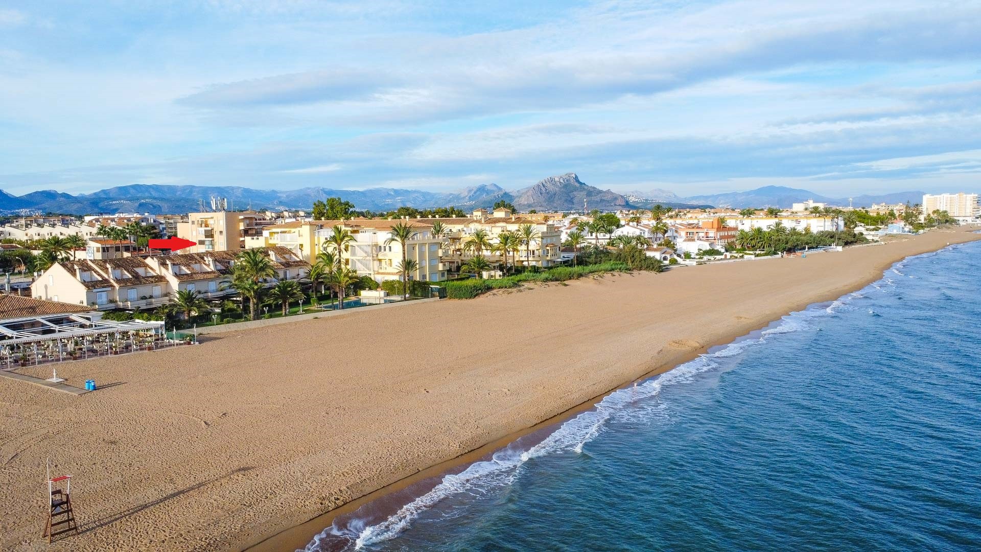 Perspectiva aérea de la amplia playa de arena y la costa mediterránea, destacando la ubicación del apartamento frente al mar y las montañas.