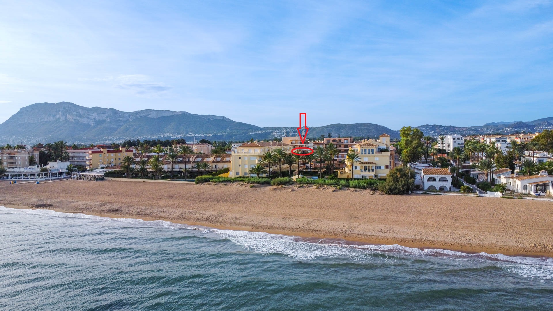 Perspectiva aérea de un complejo residencial en primera línea de playa con la vivienda señalada, ubicada frente a la arena y con vistas a la montaña.