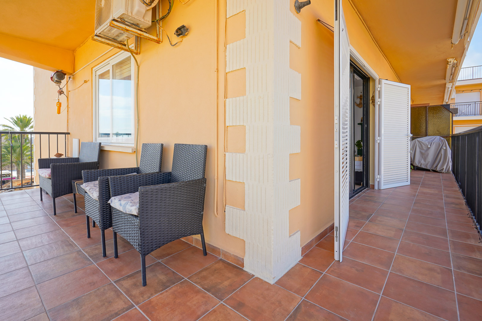 Terraza cubierta con suelos de gres rústico, sillones de mimbre oscuro y contraventanas blancas. Dispone de aire acondicionado y barandilla metálica perimetral.