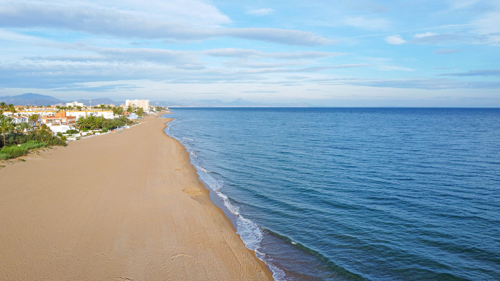 Vista aérea de una amplia playa de arena dorada junto al mar Mediterráneo, con edificaciones costeras de baja altura y montañas en el horizonte bajo un cielo despejado.