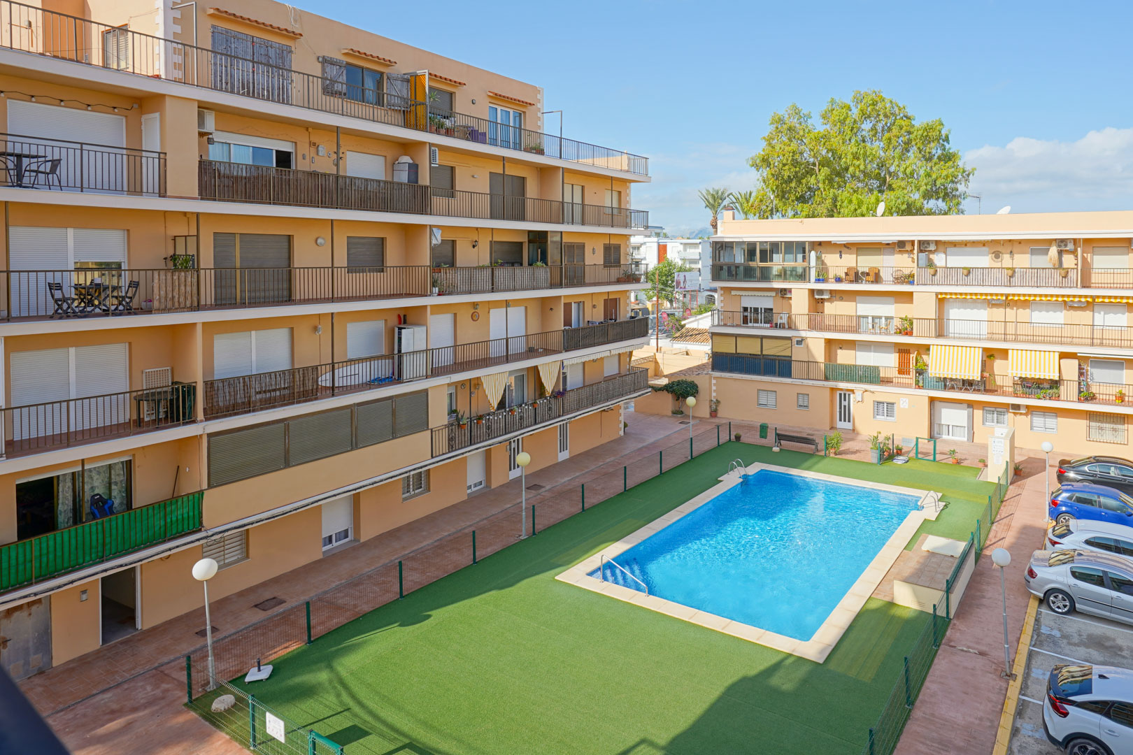 Vista del complejo residencial con piscina rectangular, zona de césped artificial vallada y edificios de varias plantas con amplios balcones y aparcamiento.