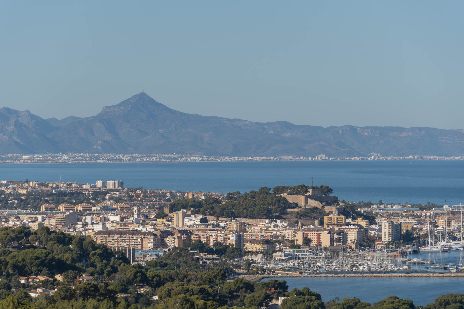 Vista panorámica de la ciudad costera con su castillo histórico, el puerto deportivo con numerosos yates y una cordillera montañosa al fondo sobre el Mediterráneo.