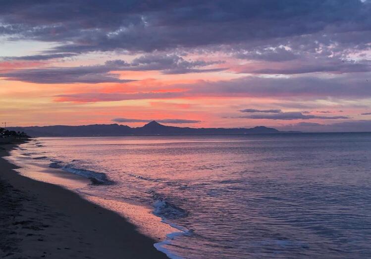 Vista panorámica de la costa arenosa al atardecer, destacando la silueta de las montañas bajo un cielo en tonos púrpuras y naranjas.
