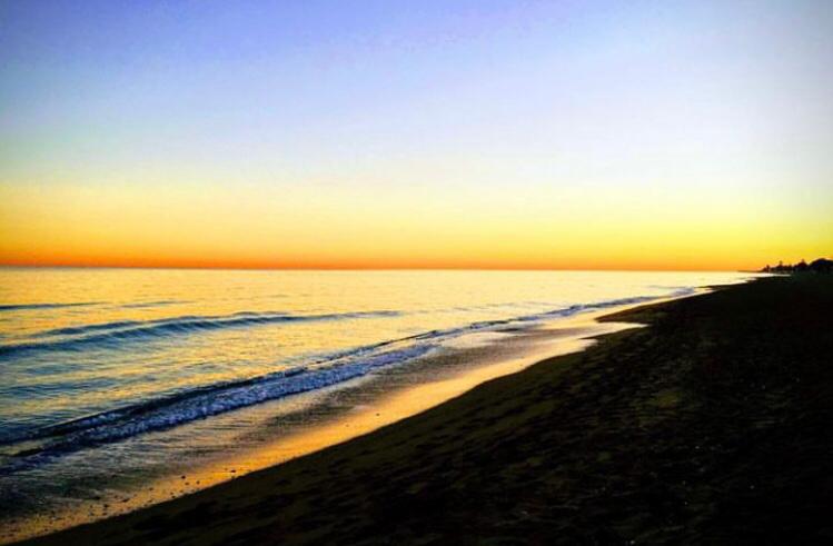 Vista panorámica de una playa de arena oscura al atardecer, con olas suaves rompiendo en la orilla bajo un cielo con degradado de azul a dorado.