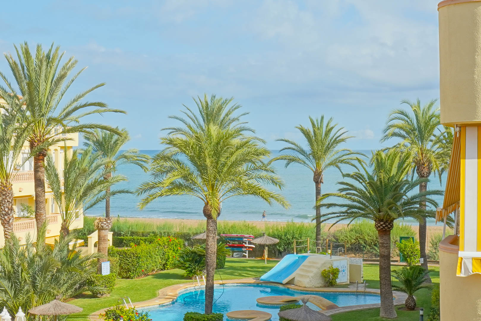 Vistas a la zona de piscina comunitaria con tobogán, rodeada de palmeras y jardines, con acceso visual directo a la playa y al mar Mediterráneo.