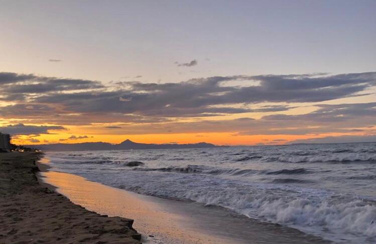 Vistas del atardecer sobre una playa de arena con olas rompiendo y siluetas de montañas bajo un cielo nublado en tonos naranja.