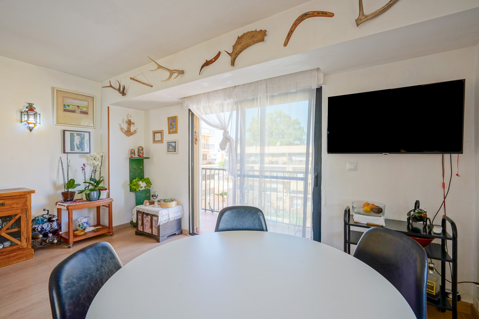 Zona de comedor con mesa redonda blanca, televisor de pared y salida a balcón. Destacan los elementos decorativos como astas y bumeranes en la viga superior.